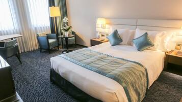 A hotel room with a large bed, a desk, a chair, a small table with a lamp, and a potted plant.