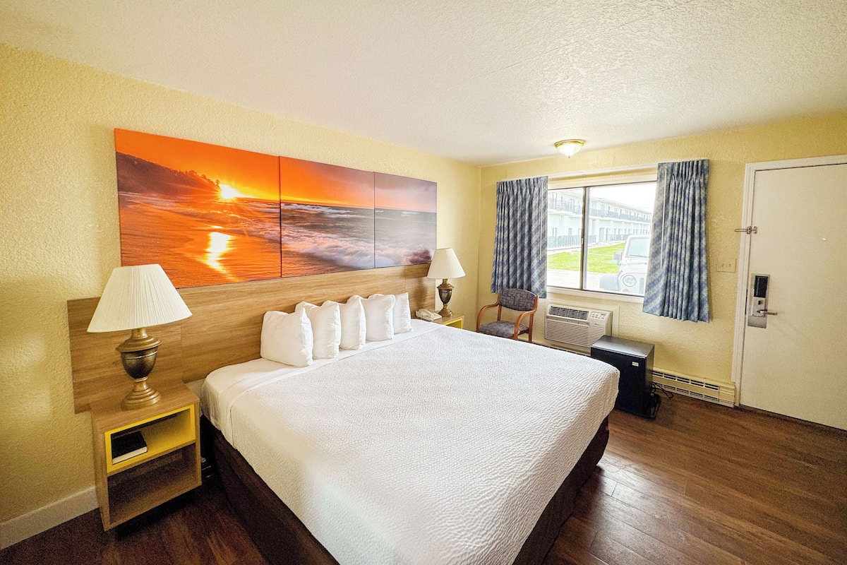 Ein Hotelzimmer mit einem großen Bett, einem Nachttisch, einer Lampe, einem Sessel, einem Fenster mit Vorhängen und einem Sonnenuntergangsbild.