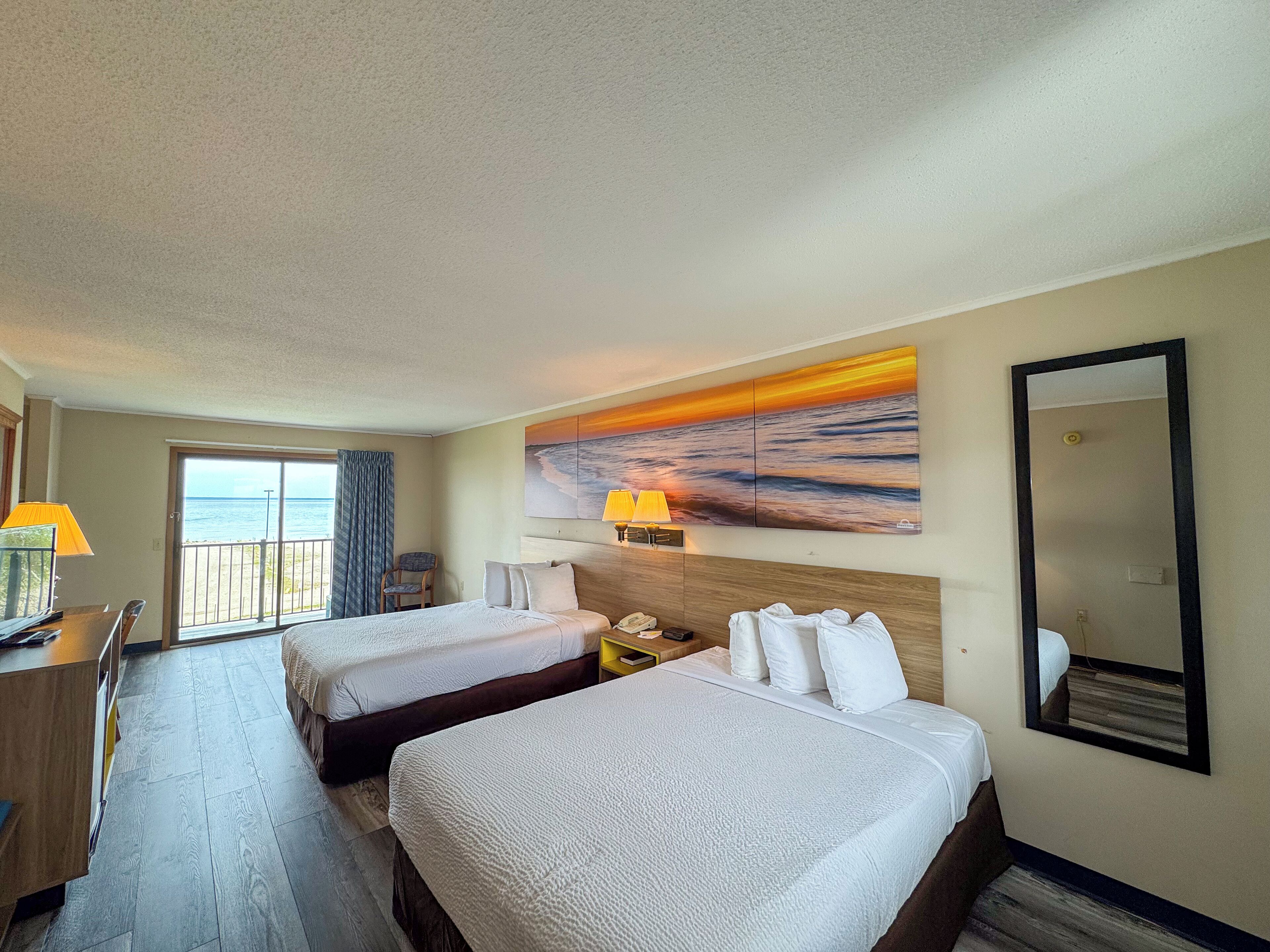 Habitación de hotel con dos camas, un escritorio, una silla, un cuadro grande de un atardecer sobre el océano y un espejo grande.