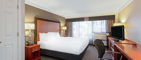 A hotel room with a large bed, a desk with a television, a chair, a nightstand with a phone, and a window with curtains.