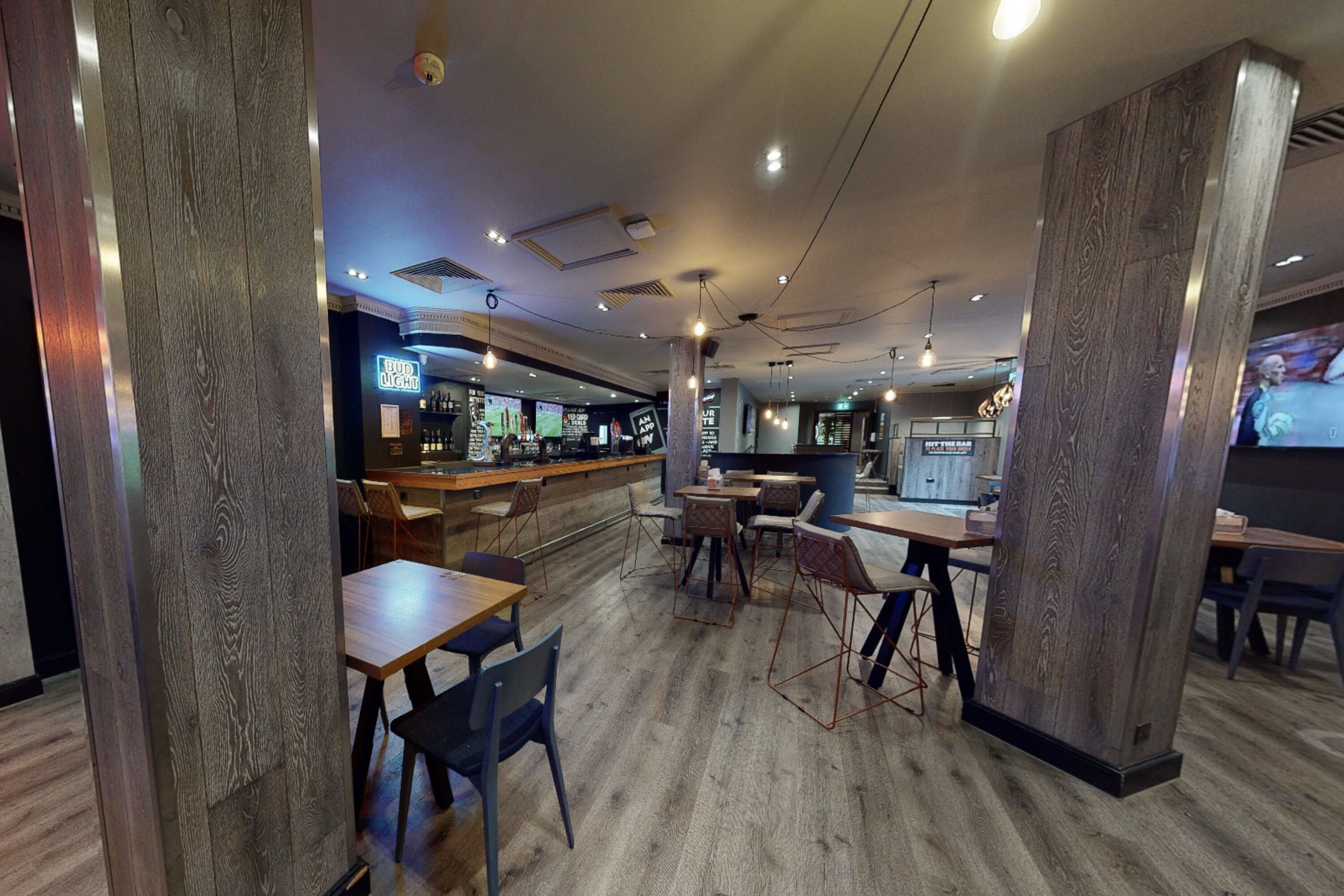 A restaurant with wooden flooring, tables, chairs, and a bar area with a neon sign.