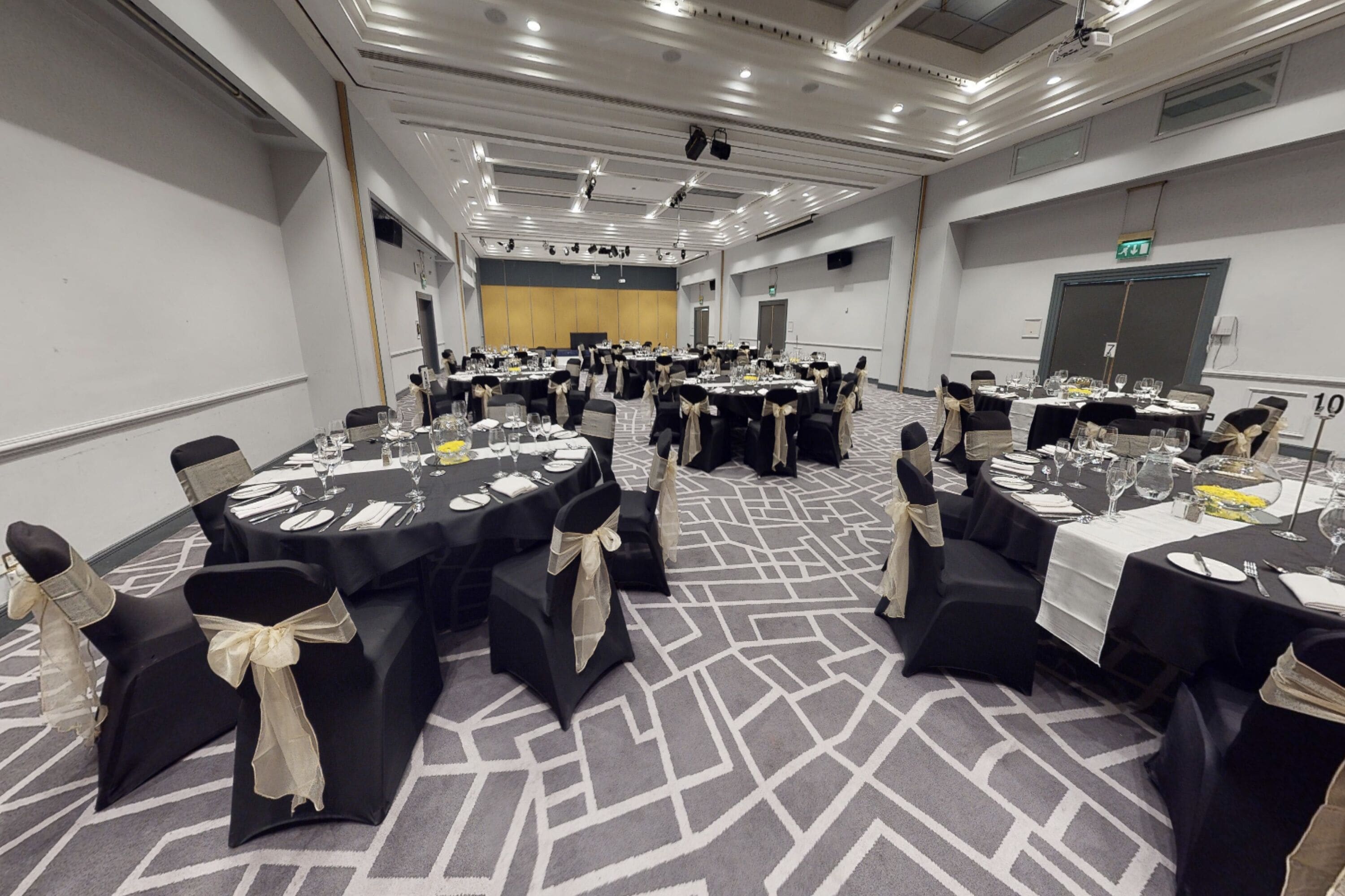 A conference room with multiple round tables, black chairs with beige sashes, and a carpeted floor.