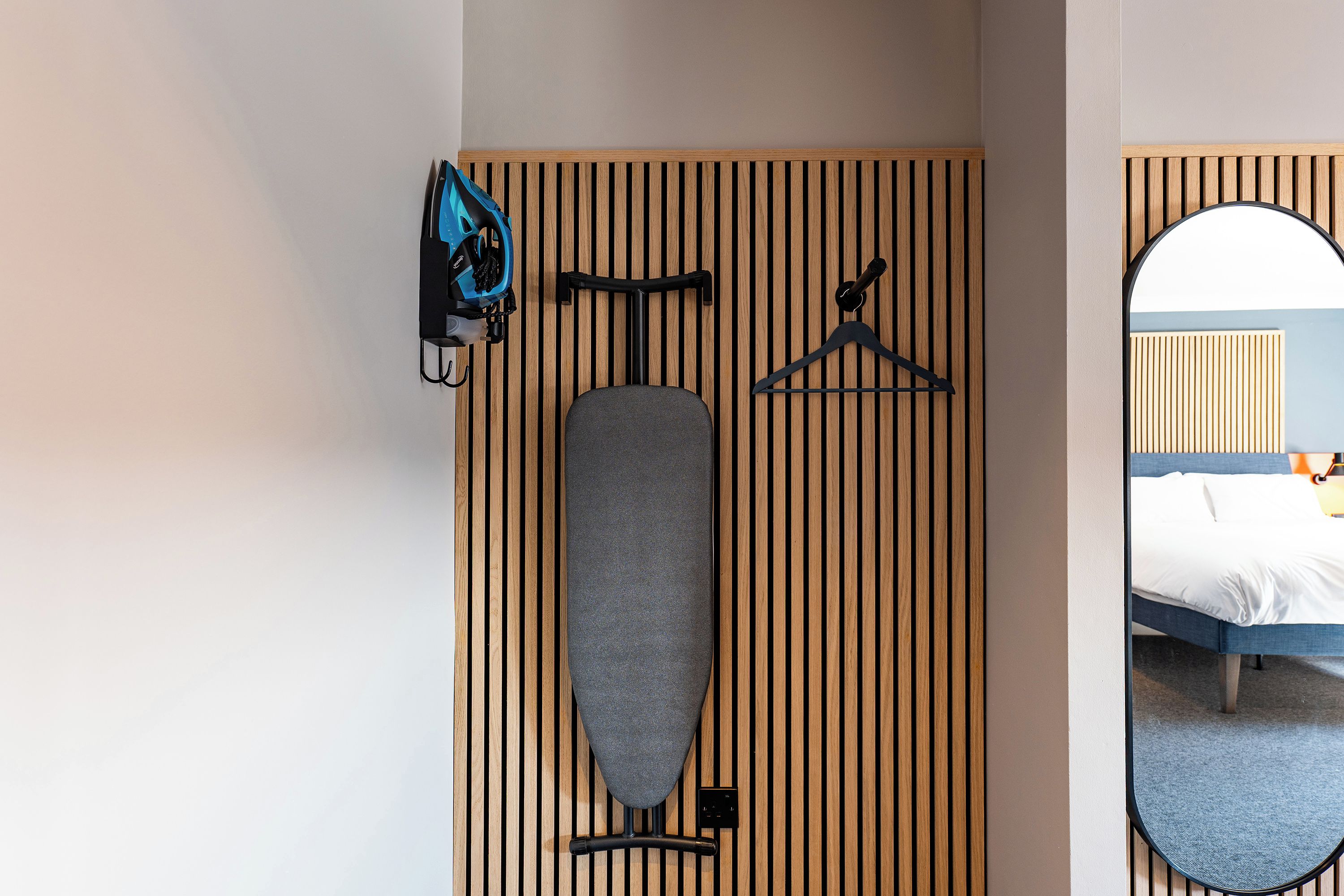 A hotel room with a wooden slat wall, a bed, a mirror, and a hanging rack with a blue bag and a hanger.