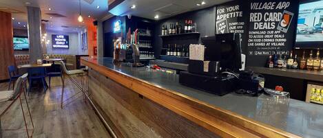A bar area with a wooden counter, bar stools, and a neon sign. There are shelves with bottles and glasses, and a sign promoting a Village App for deals.