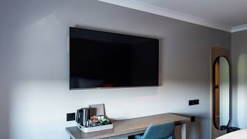 A hotel room with a flat-screen TV mounted on the wall, a wooden desk with a chair, a small tray of toiletries, and a mirror on the wall.