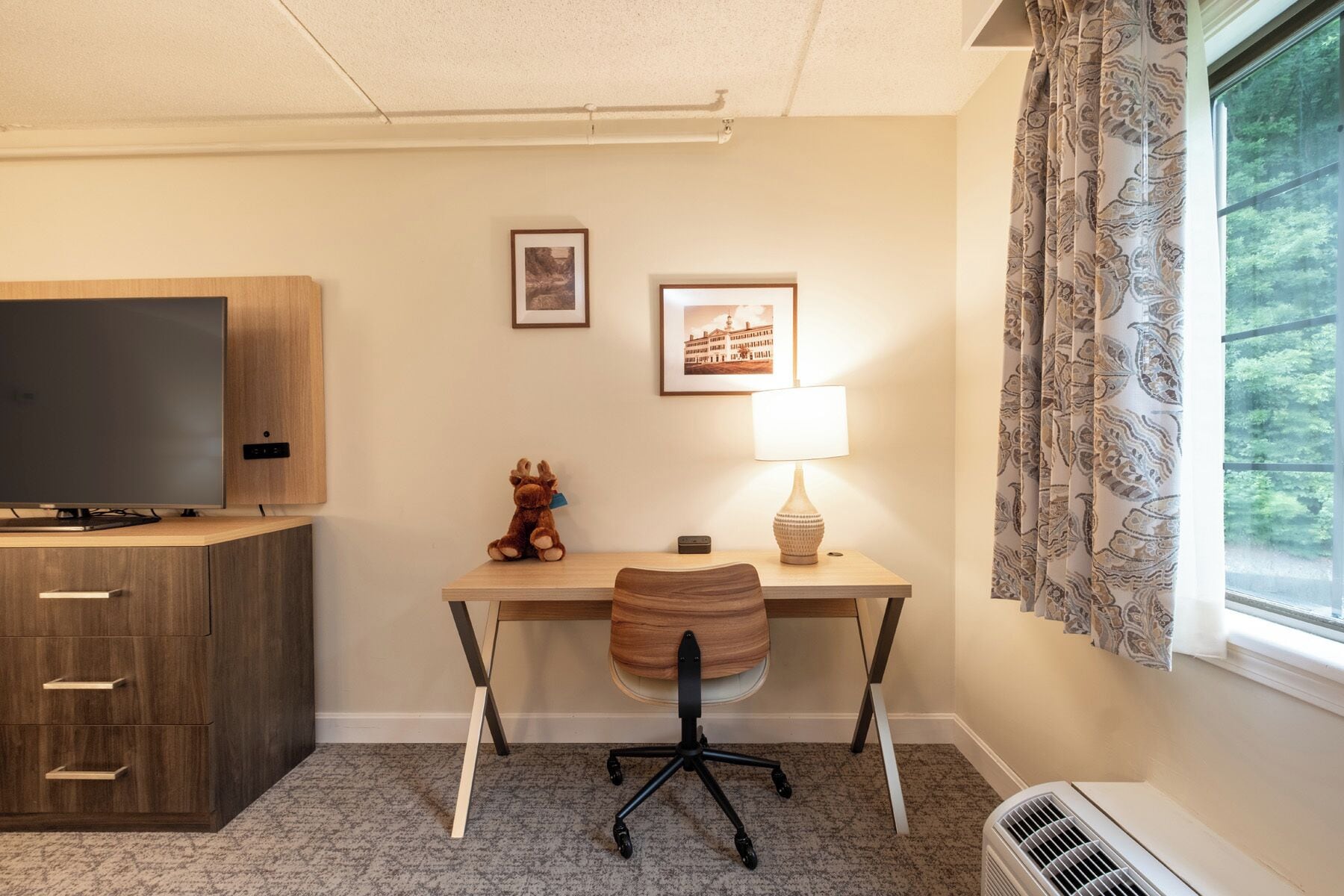 A hotel room with a wooden desk, a television, a lamp, a chair, a small statue, and a window with curtains.