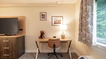 A hotel room with a wooden desk, a television, a lamp, a chair, a small statue, and a window with curtains.