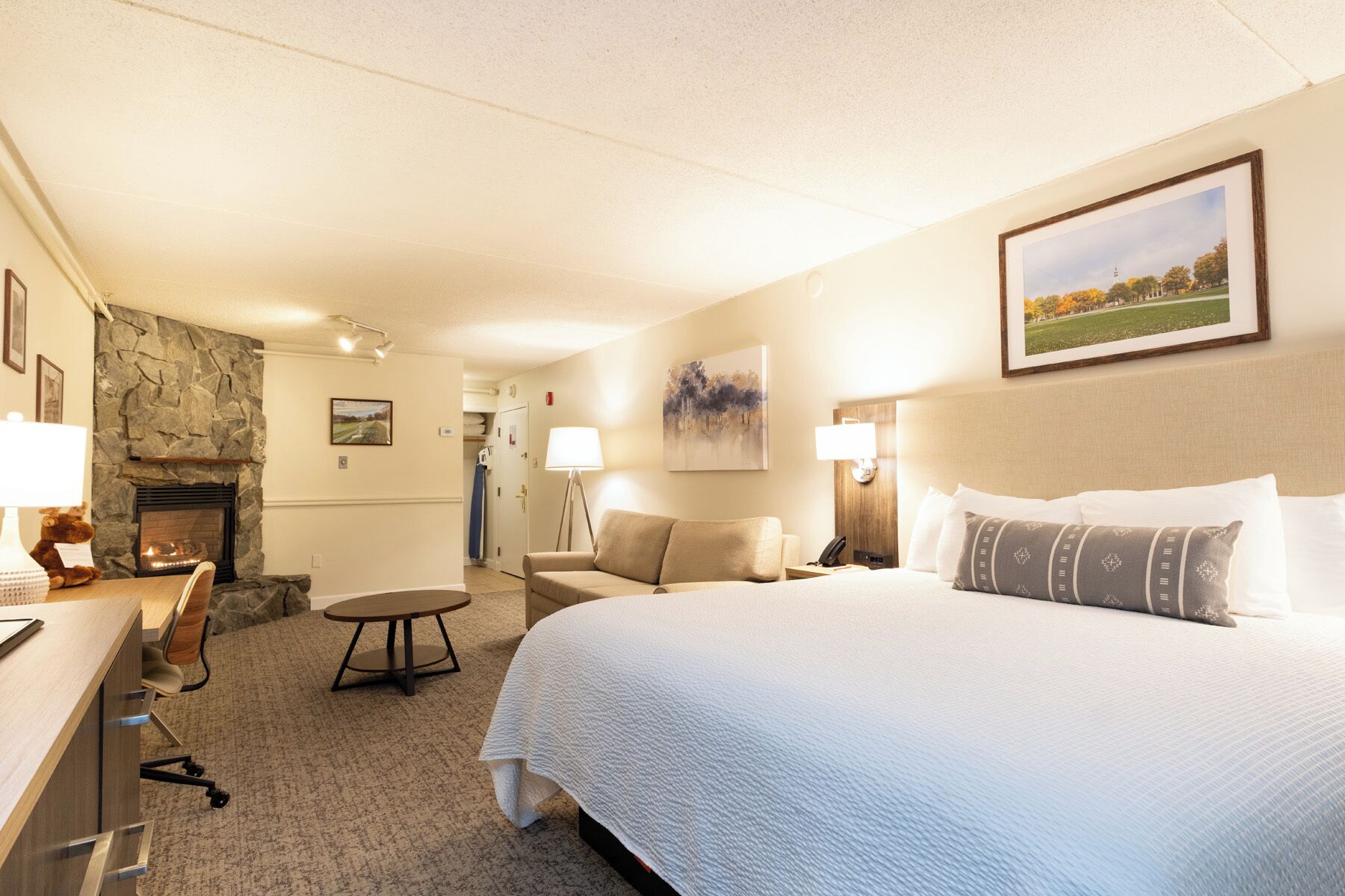 A hotel room with a bed, a desk, a chair, a fireplace, and a painting on the wall.