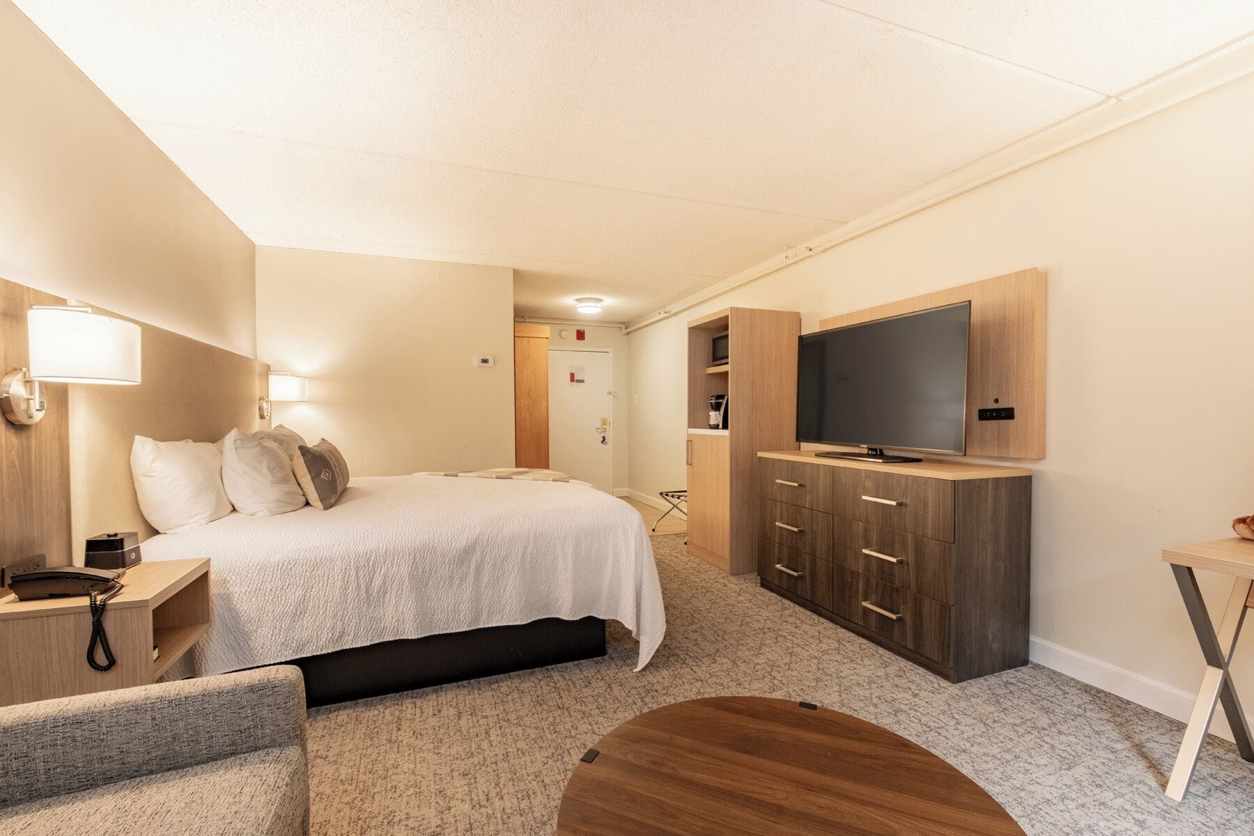 Een hotelkamer met een bed, nachtkastje, lamp, televisie, kledingkast en een klein rond tafeltje.