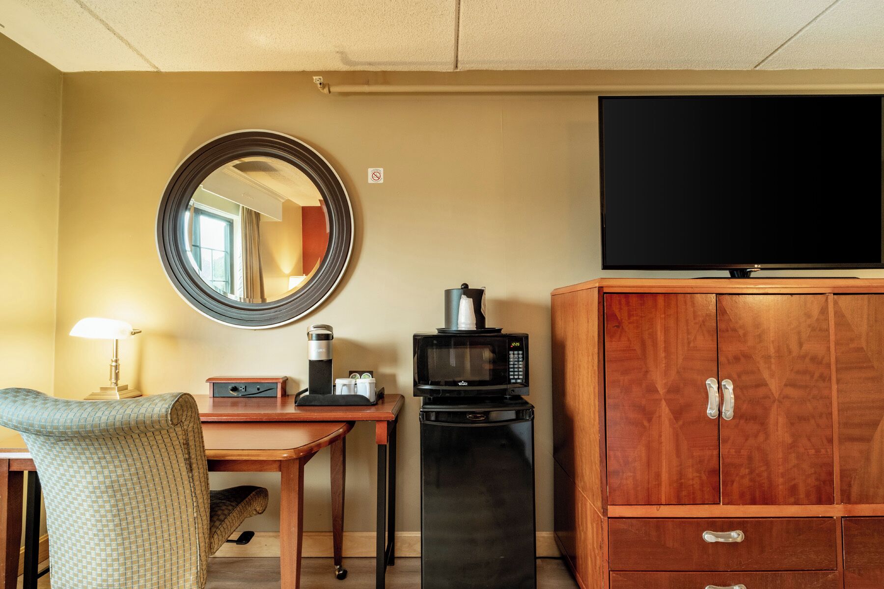 Een hotelkamer met een houten kast, een televisie, een magnetron, een koffiezetapparaat, een bureau met een lamp en een stoel.