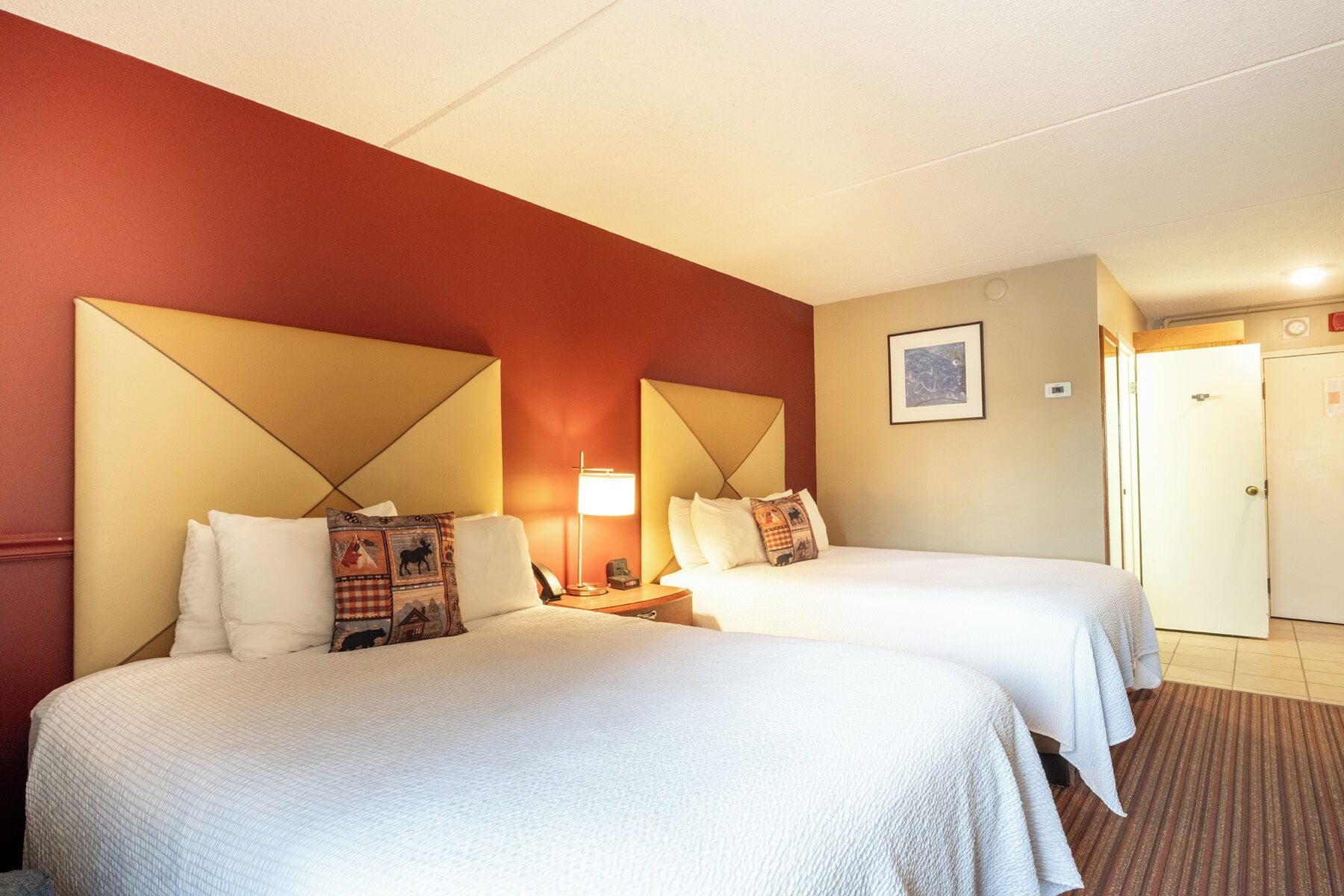 A hotel room with two beds, a nightstand with a lamp, a framed picture on the wall, and a door in the background.