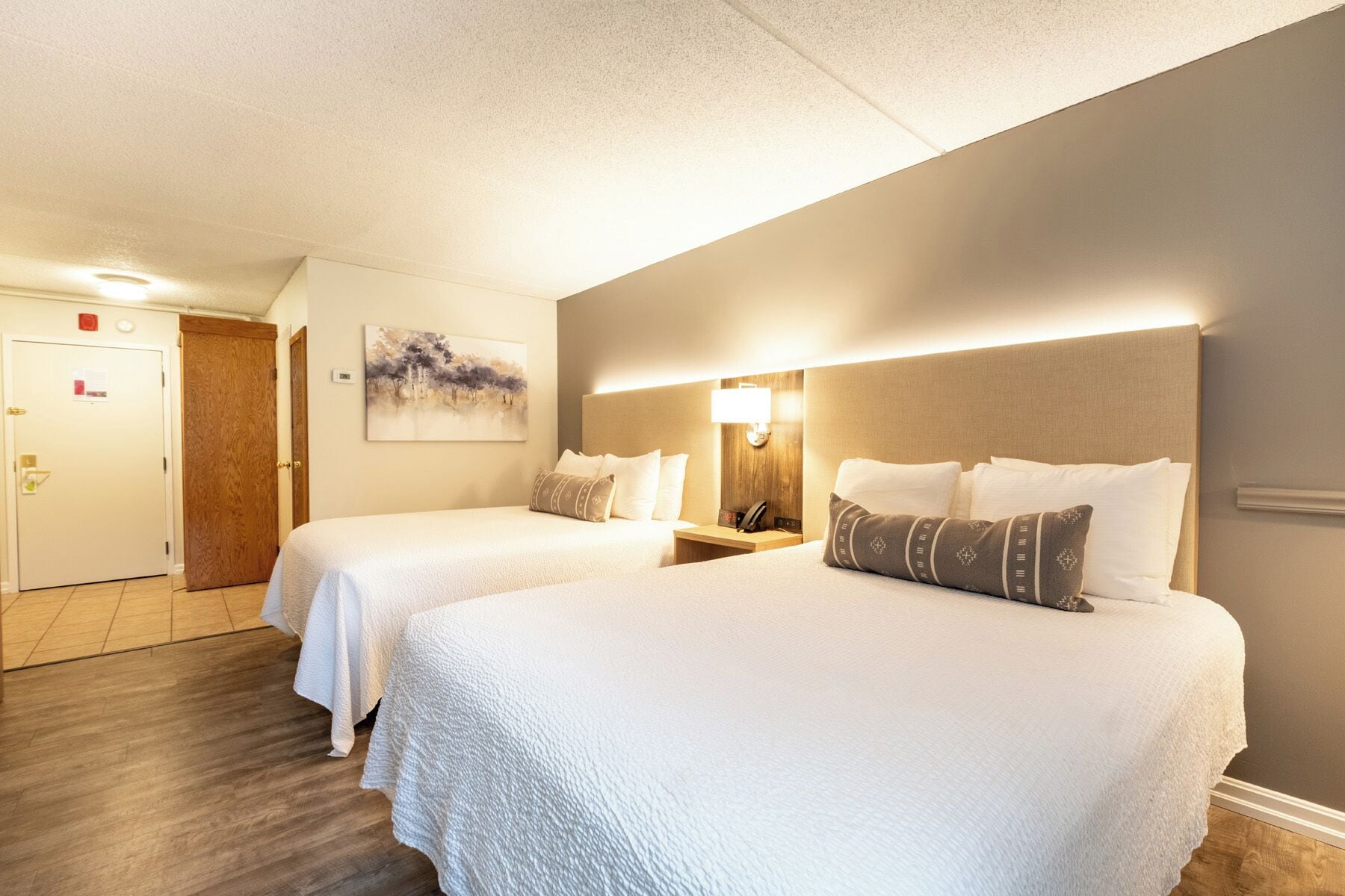 A hotel room with two beds, a painting on the wall, a bedside table with a lamp, and a door with a red sign.