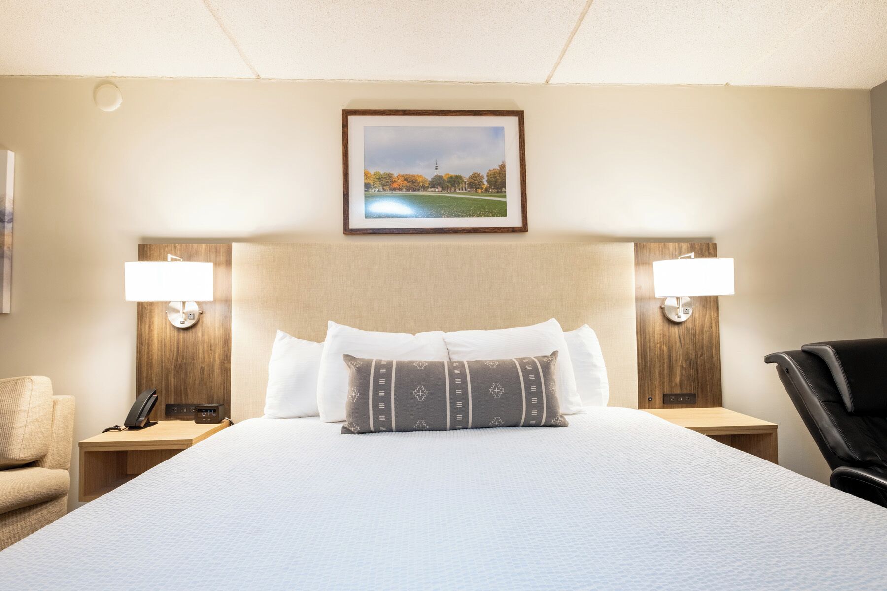 A hotel room with a large bed, two bedside lamps, a wooden headboard, a framed picture on the wall, and a desk with a phone.