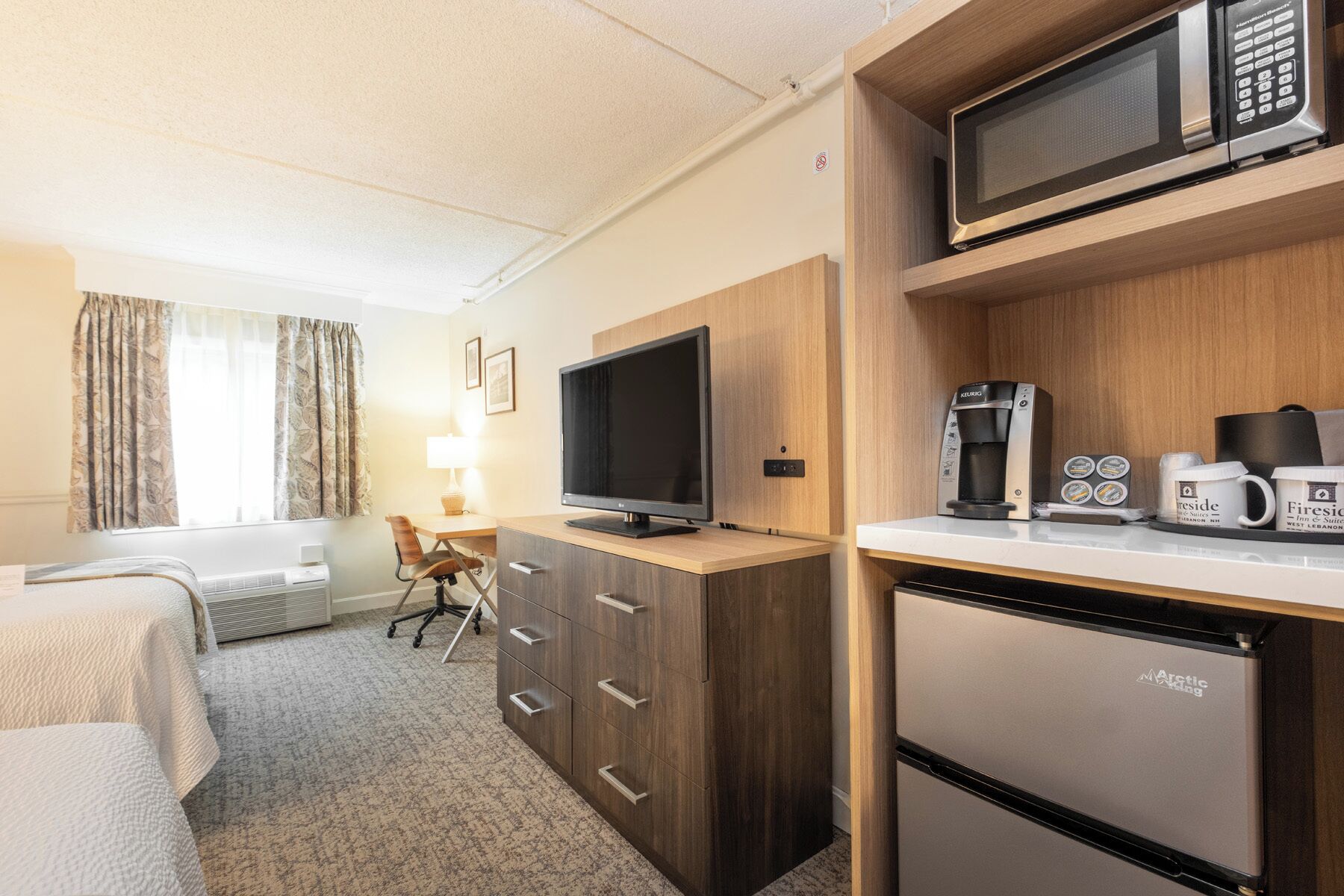 Habitación de hotel con cama, escritorio, televisor, cafetera y un refrigerador pequeño.