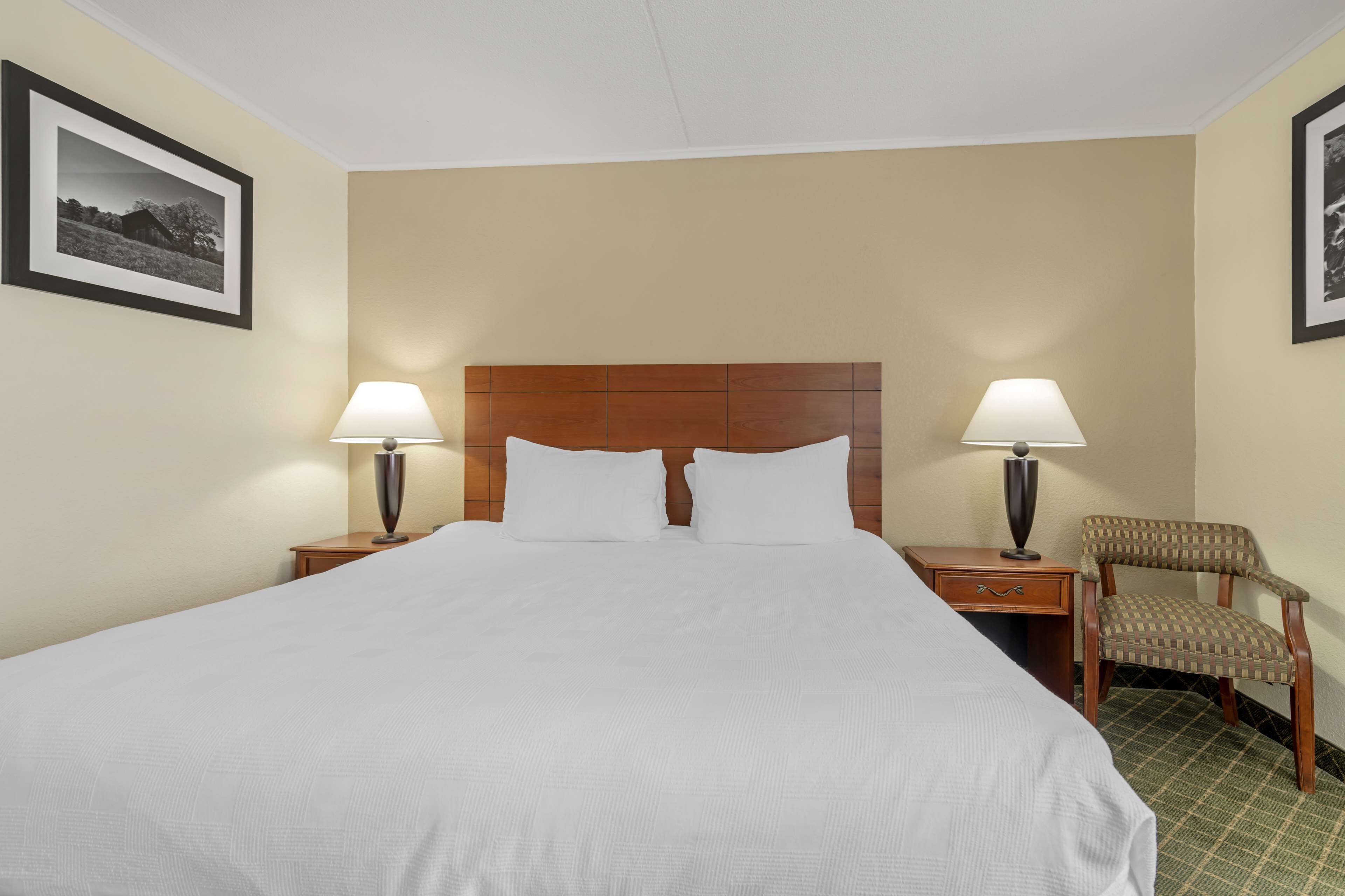 A hotel room with a large bed, two bedside lamps, a wooden headboard, a chair, and two framed pictures on the walls.