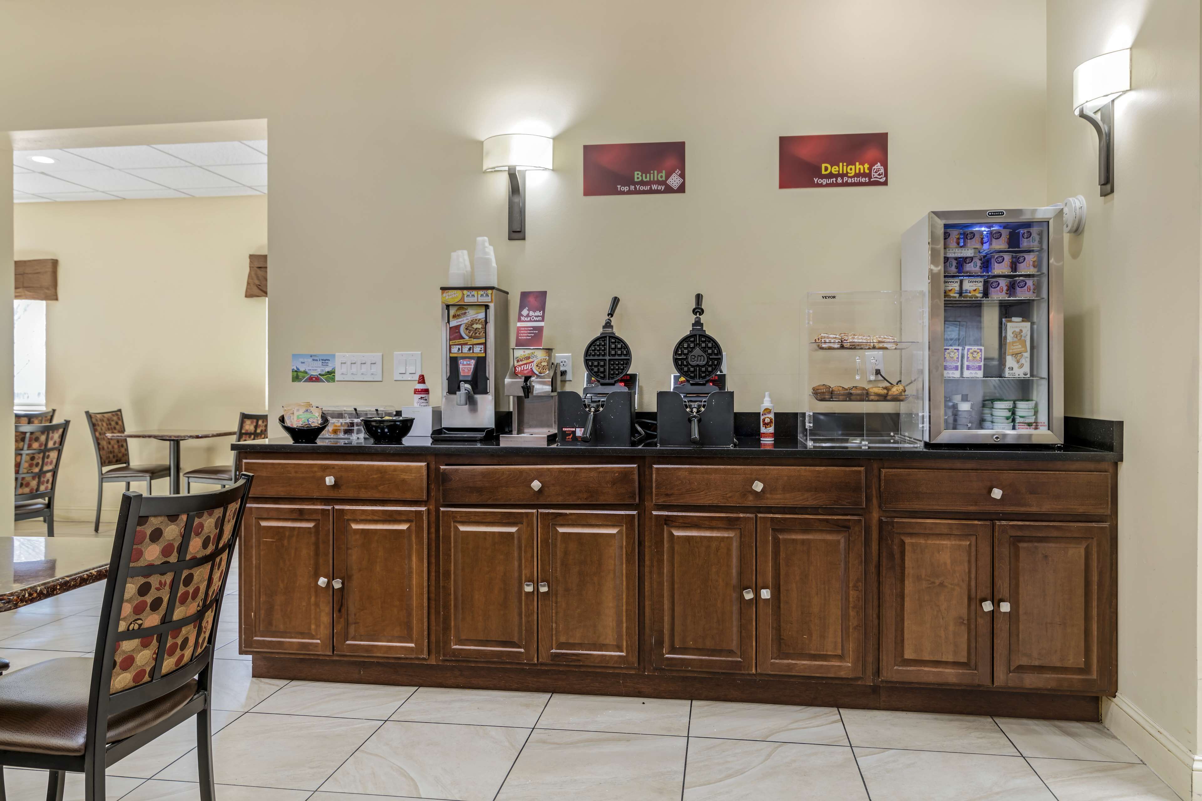 A hotel reception area with a wooden counter, a coffee machine, a waffle maker, and a refrigerator stocked with beverages.