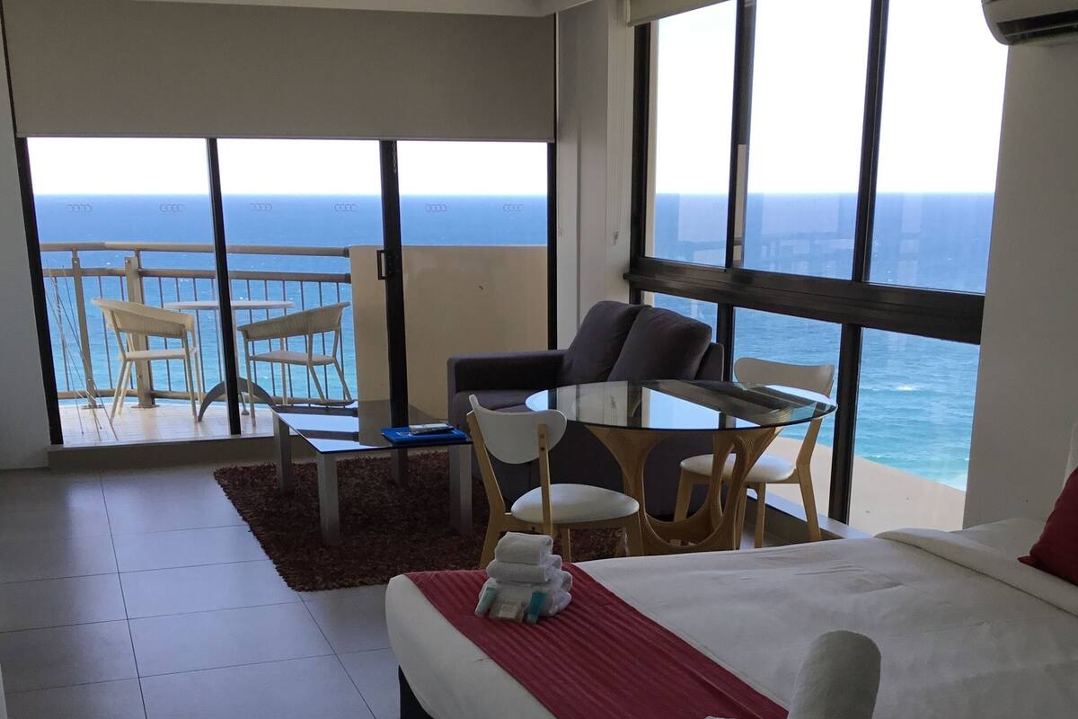 A hotel room with a large bed, a balcony with a view of the ocean, a dining table, and two chairs.