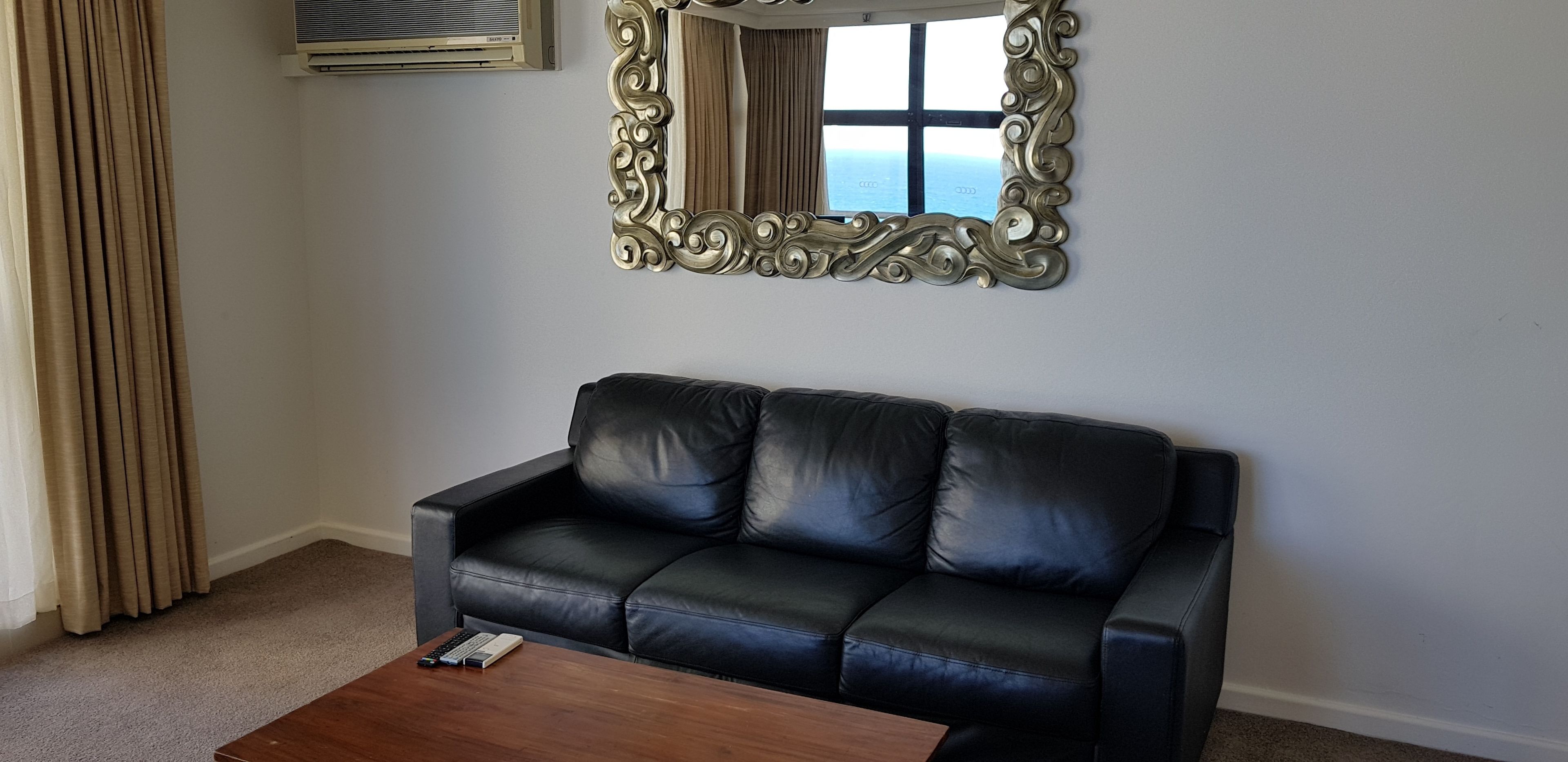 Ein schwarzes Ledersofa an einer weißen Wand, ein Couchtisch aus Holz davor und ein großer, verzierter Spiegel darüber.