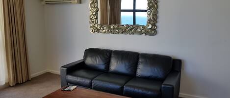 Ein schwarzes Ledersofa an einer weißen Wand, ein Couchtisch aus Holz davor und ein großer, verzierter Spiegel darüber.