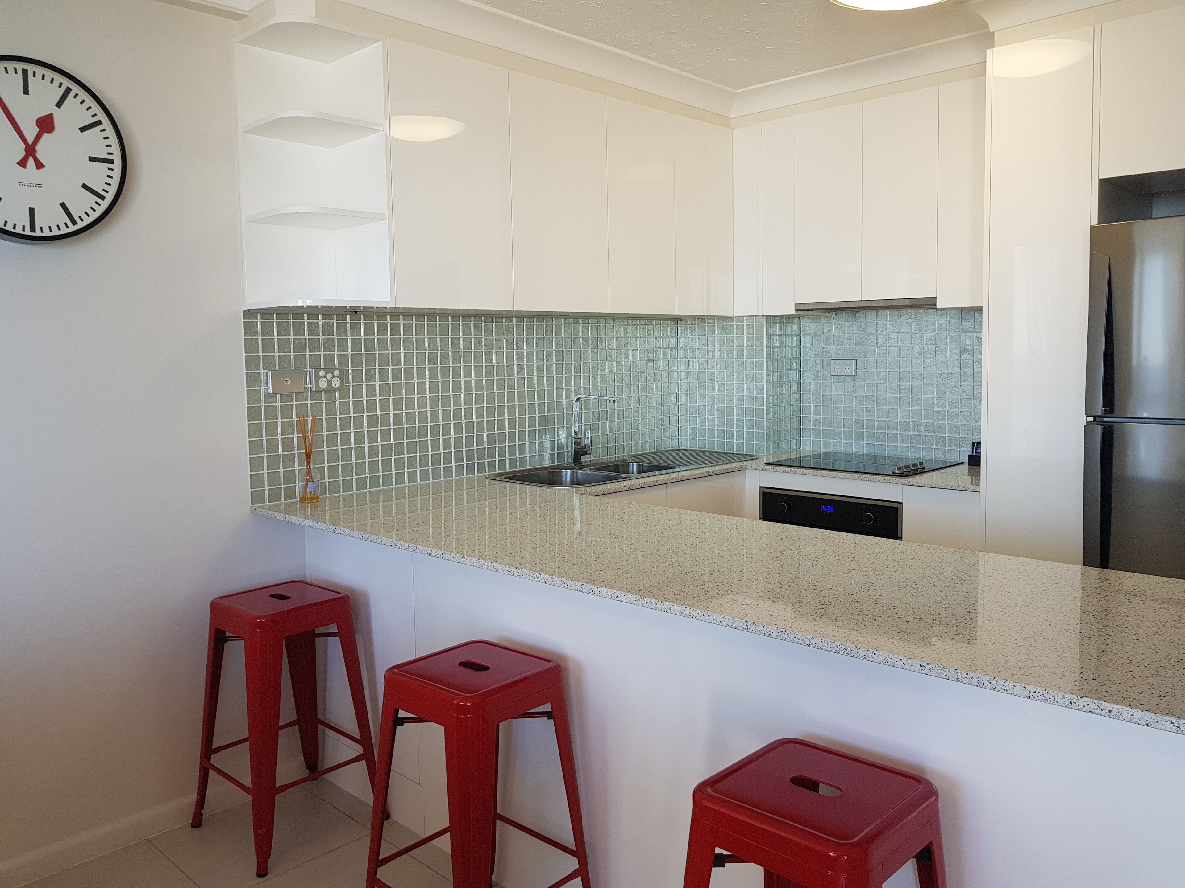 A modern kitchen with white cabinets, a stainless steel refrigerator, a clock on the wall, and two red stools.