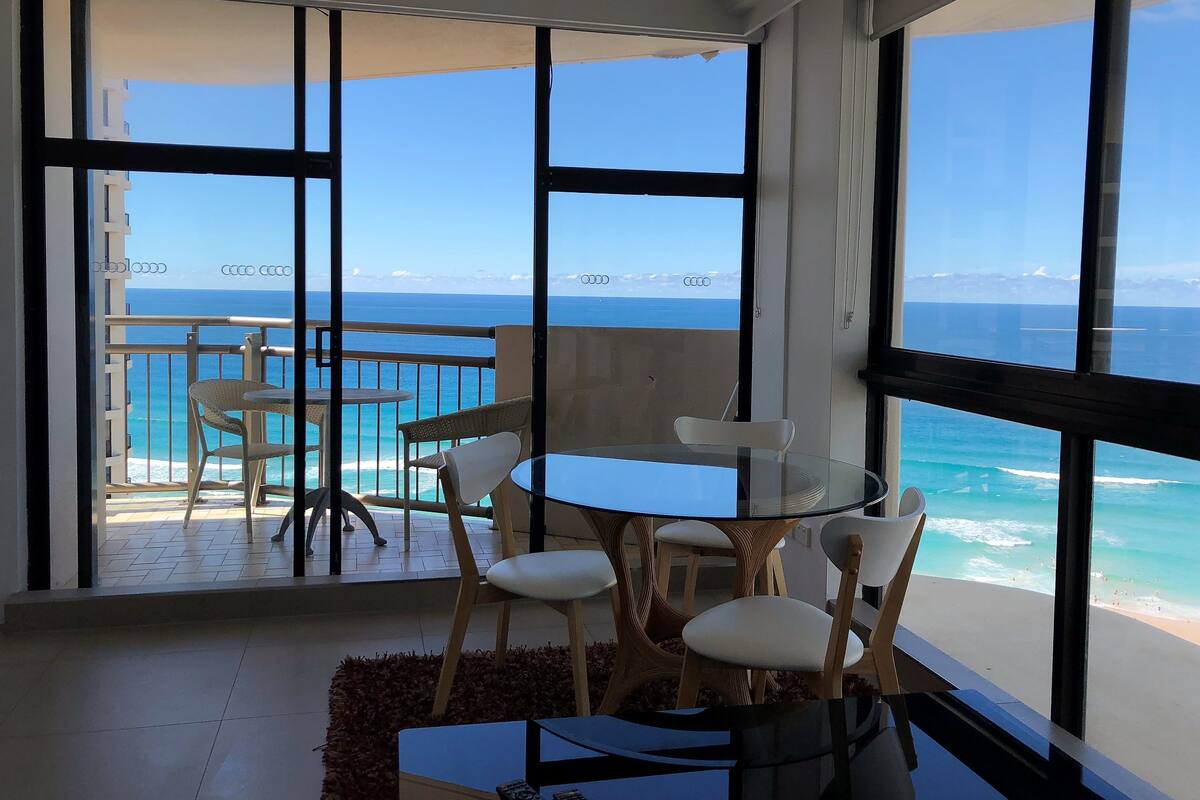 A balcony with a glass table and chairs, overlooking a beach and ocean.