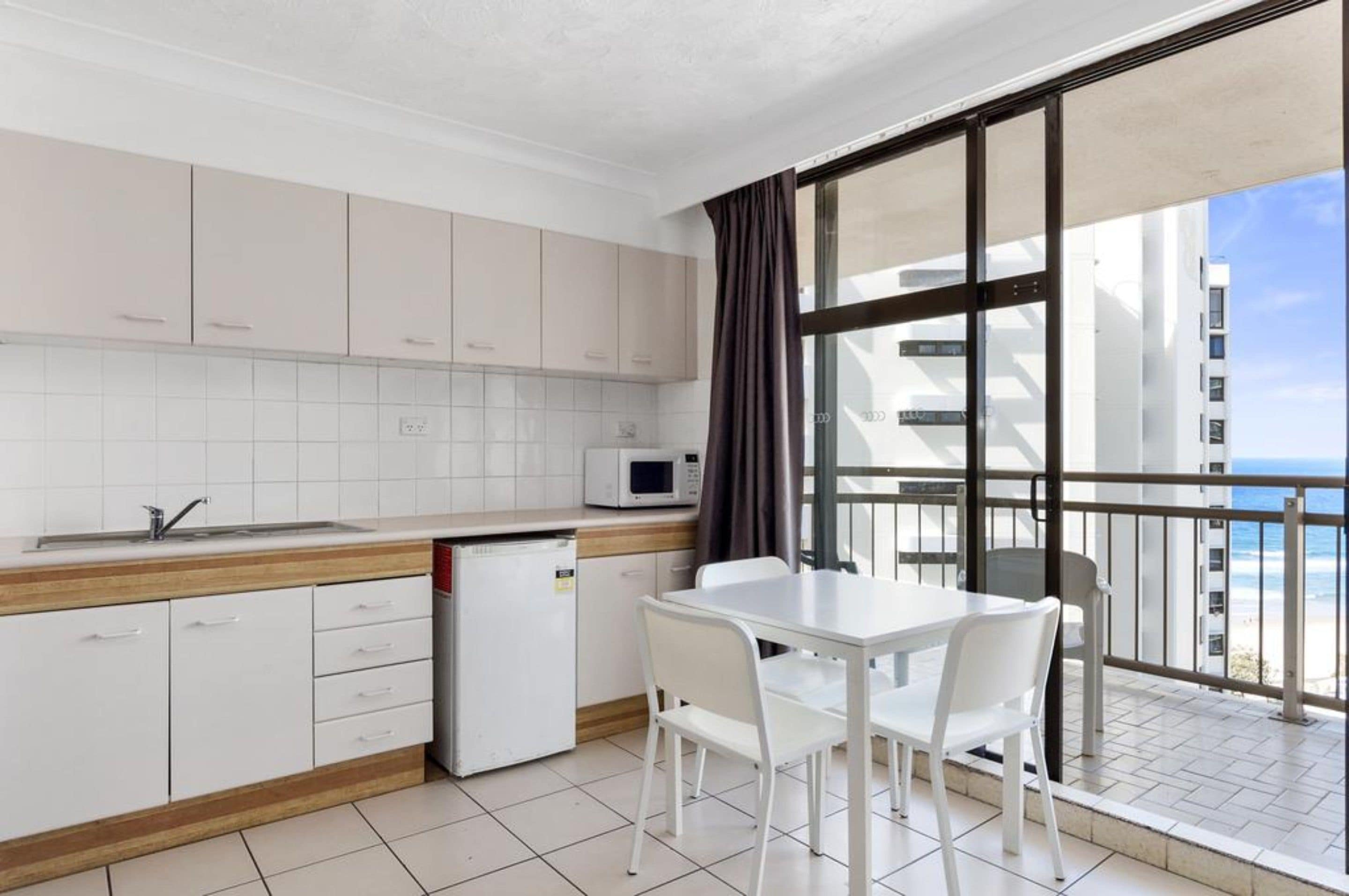 A modern kitchen with white cabinets, a microwave, and a small refrigerator. There is a dining area with a white table and chairs, and a balcony with a view of the ocean and buildings.