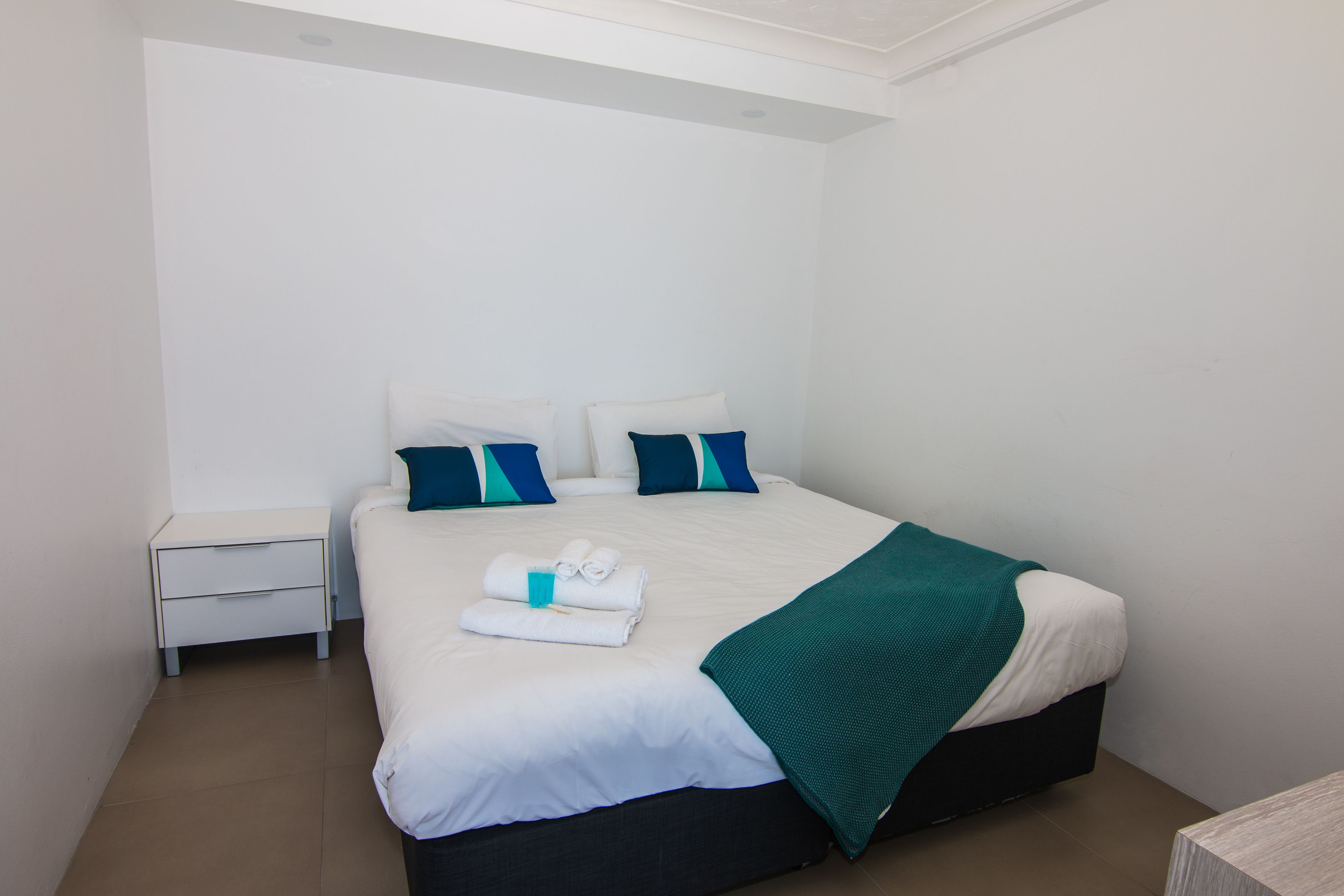 A neatly made bed with white bedding and a teal throw blanket, a white bedside table, and a dark-colored headboard.