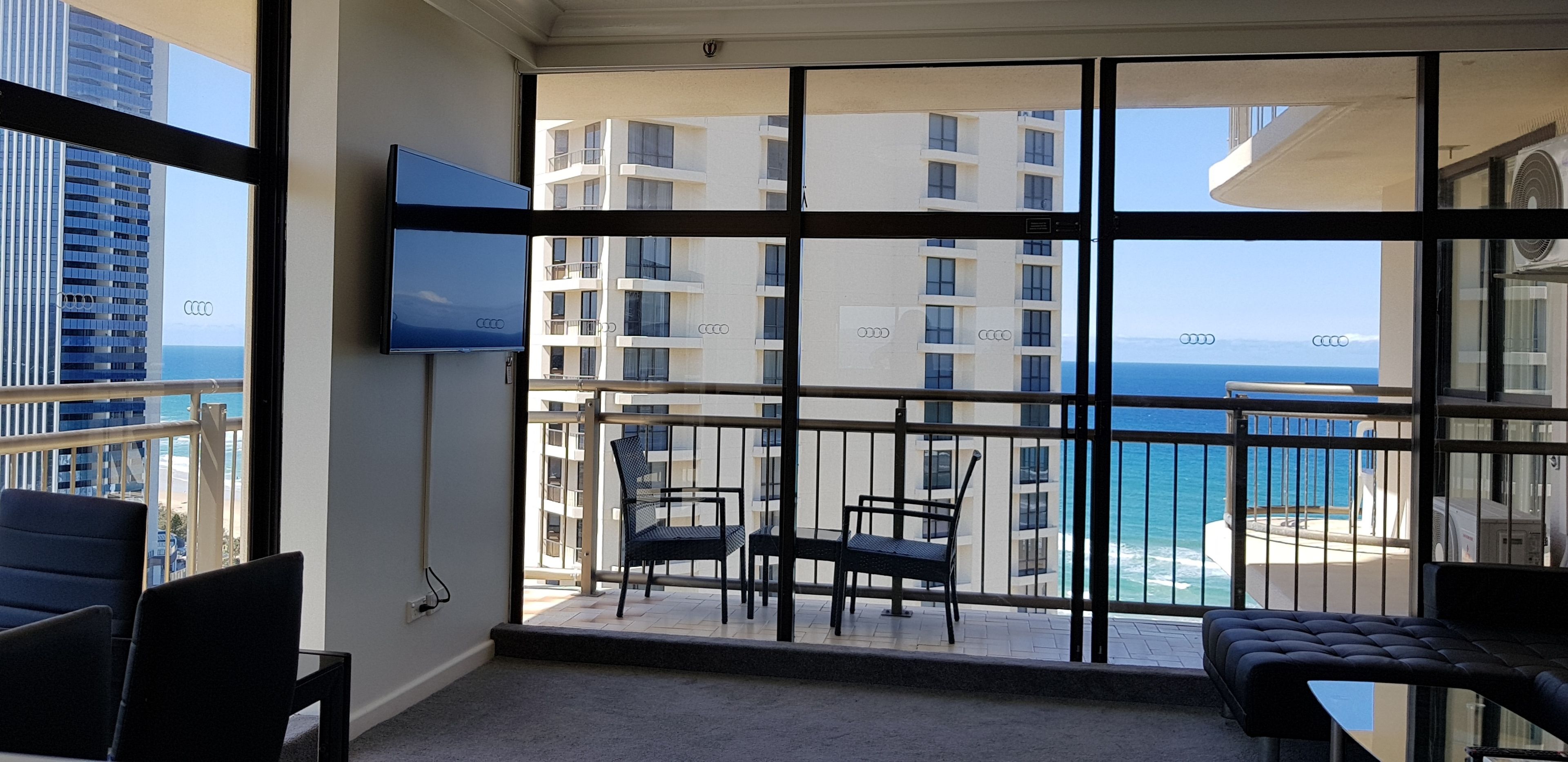 A balcony with a view of the ocean and a building across the water. There is a TV mounted on the wall and a seating area with a table and chairs.