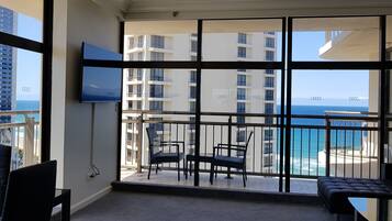 A balcony with a view of the ocean and a building across the water. There is a TV mounted on the wall and a seating area with a table and chairs.