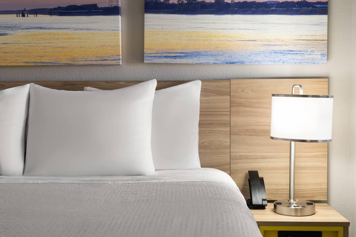 Una cama bien hecha con sábanas blancas, cabecera de madera, mesita de noche con lámpara y dos cuadros de un paisaje en la pared.