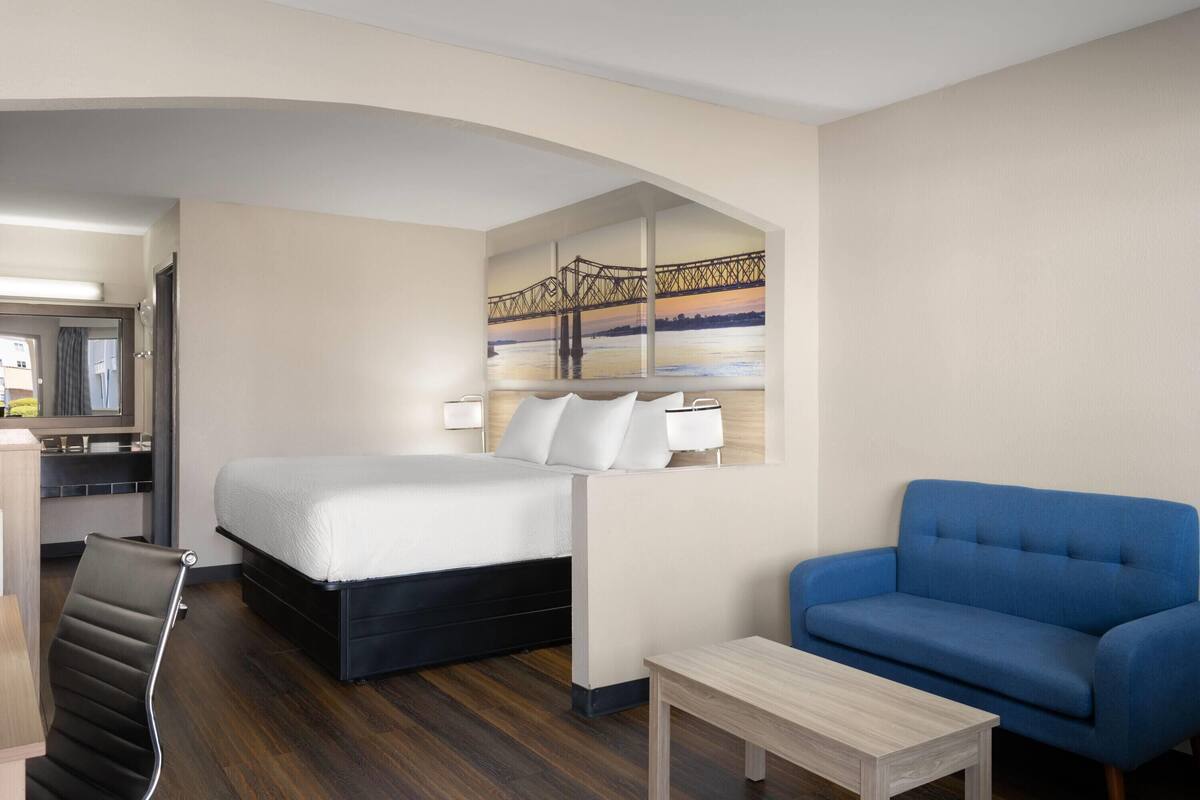 Una habitación de hotel con una cama, un sofá azul, una mesita y un cuadro de un puente en la pared.