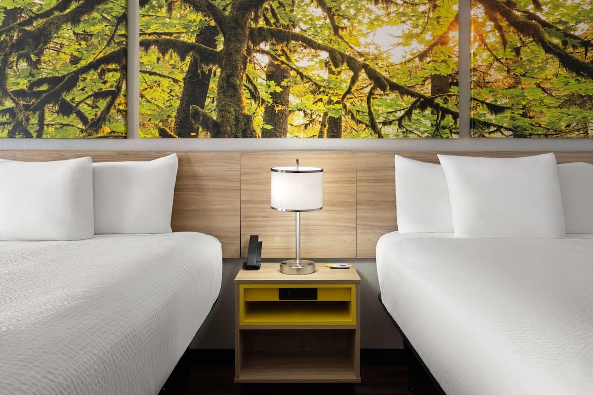 Habitación de hotel con dos camas, una mesita de noche con lámpara y un mural en la pared con un bosque.