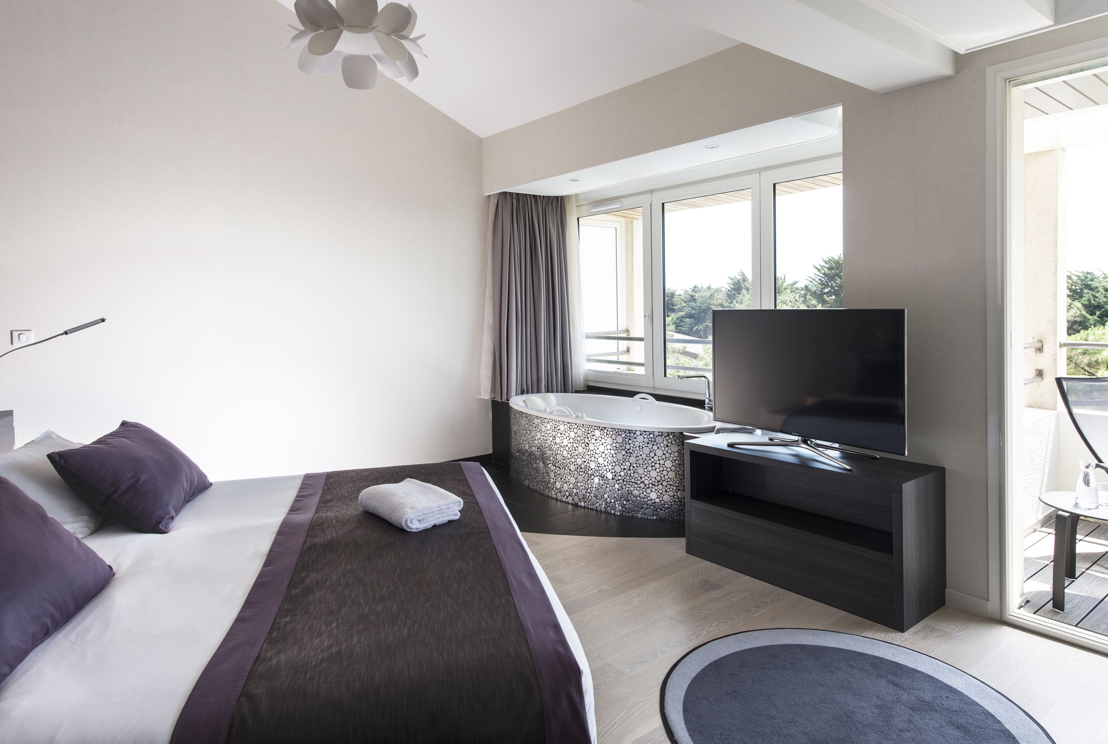 A modern hotel room with a large bed, a flat-screen TV, a bathtub, and a balcony with a view of the outdoors.