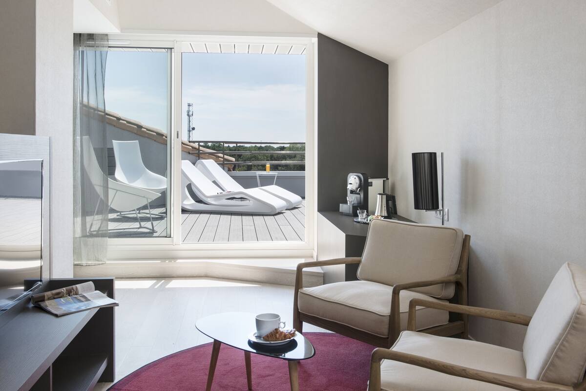 Ein modernes Hotelzimmer mit Balkon, einer Sitzecke mit zwei Stühlen, einem kleinen Tisch mit Tasse und Untertasse sowie einem Fernseher an der Wand.