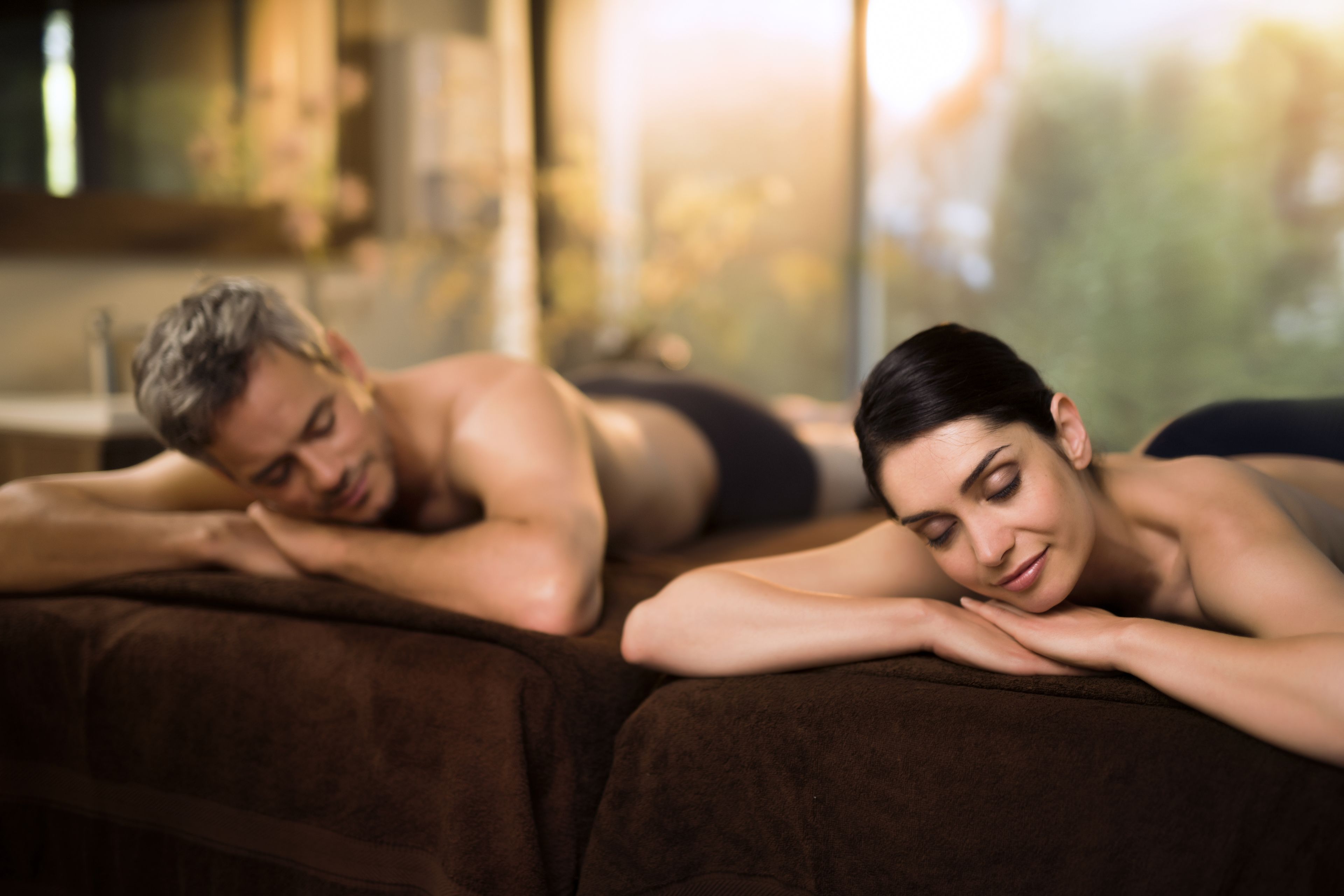 Two people lying on a massage table in a room with a large window.