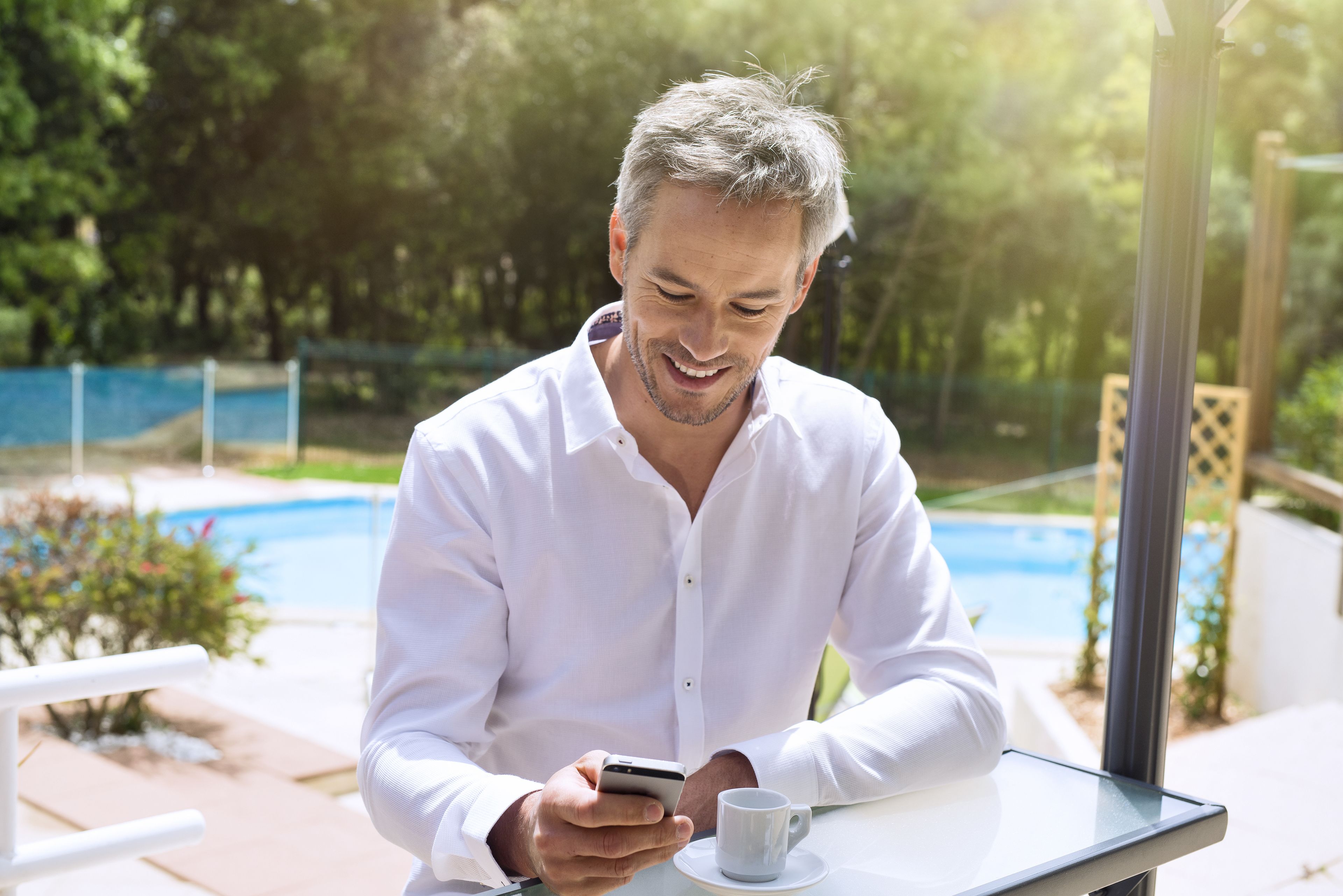 Un hombre sentado en una mesa al aire libre, con un teléfono inteligente y una taza, cerca de una piscina y vegetación.