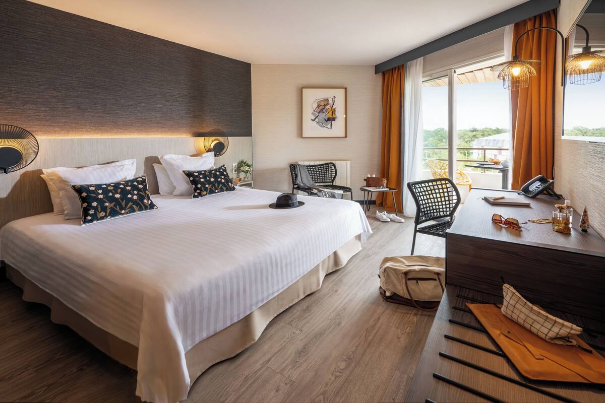 Ein Hotelzimmer mit einem großen Bett, einem Schreibtisch mit Stuhl, einem kleinen Tisch und einem Fenster mit Vorhängen.