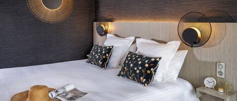 A neatly made bed with white linens, a straw hat, and a magazine on the bed. There are two wall-mounted fans and a small shelf with a clock and a bottle.