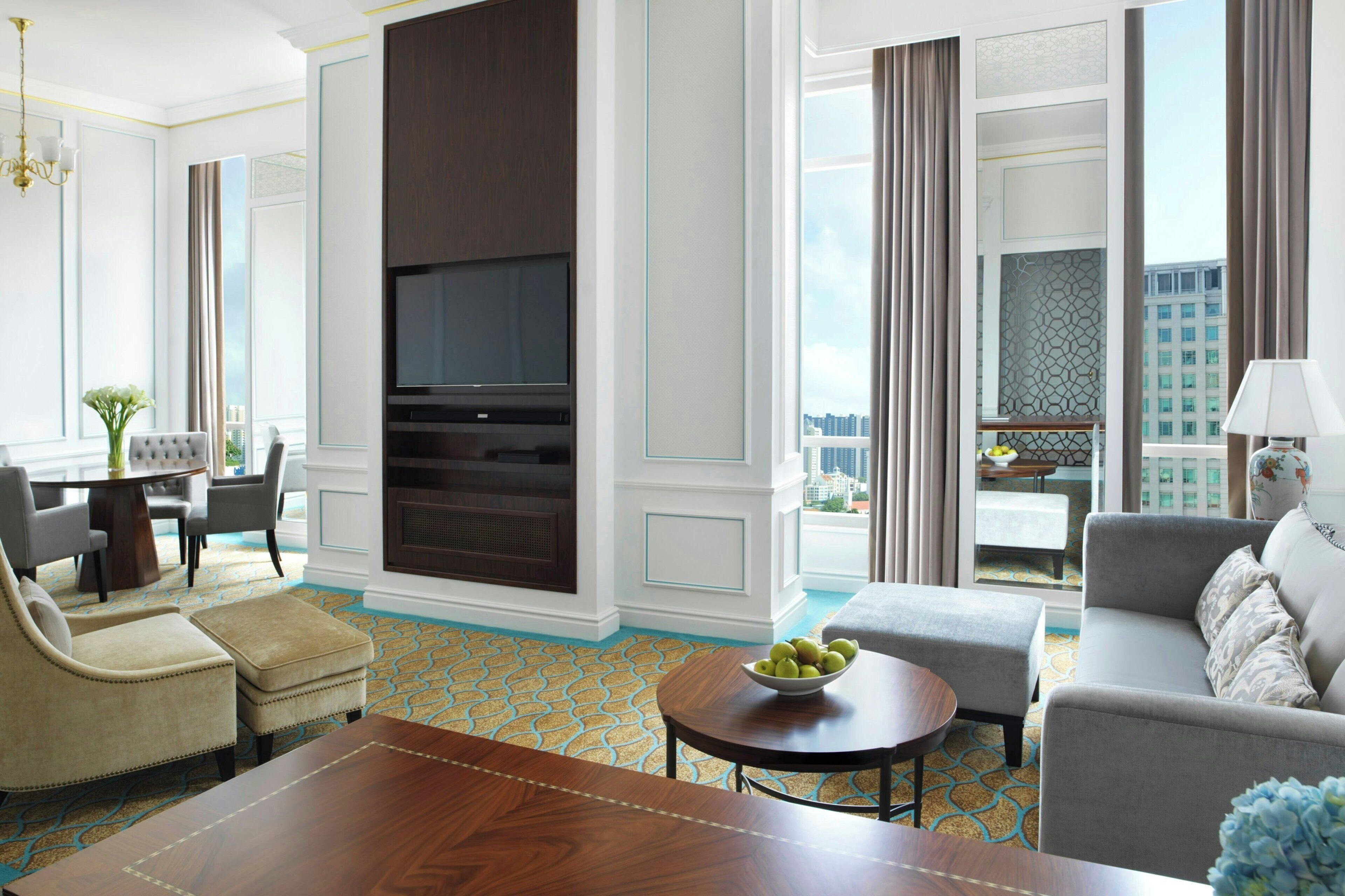 A modern hotel room with a large flat-screen TV, a dining area with a round table and chairs, a sofa, and a view of the cityscape.