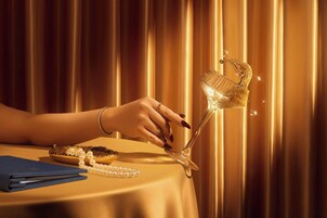 A hand holding a crystal glass with a stem, placed on a table with a blue folder and a pearl necklace.