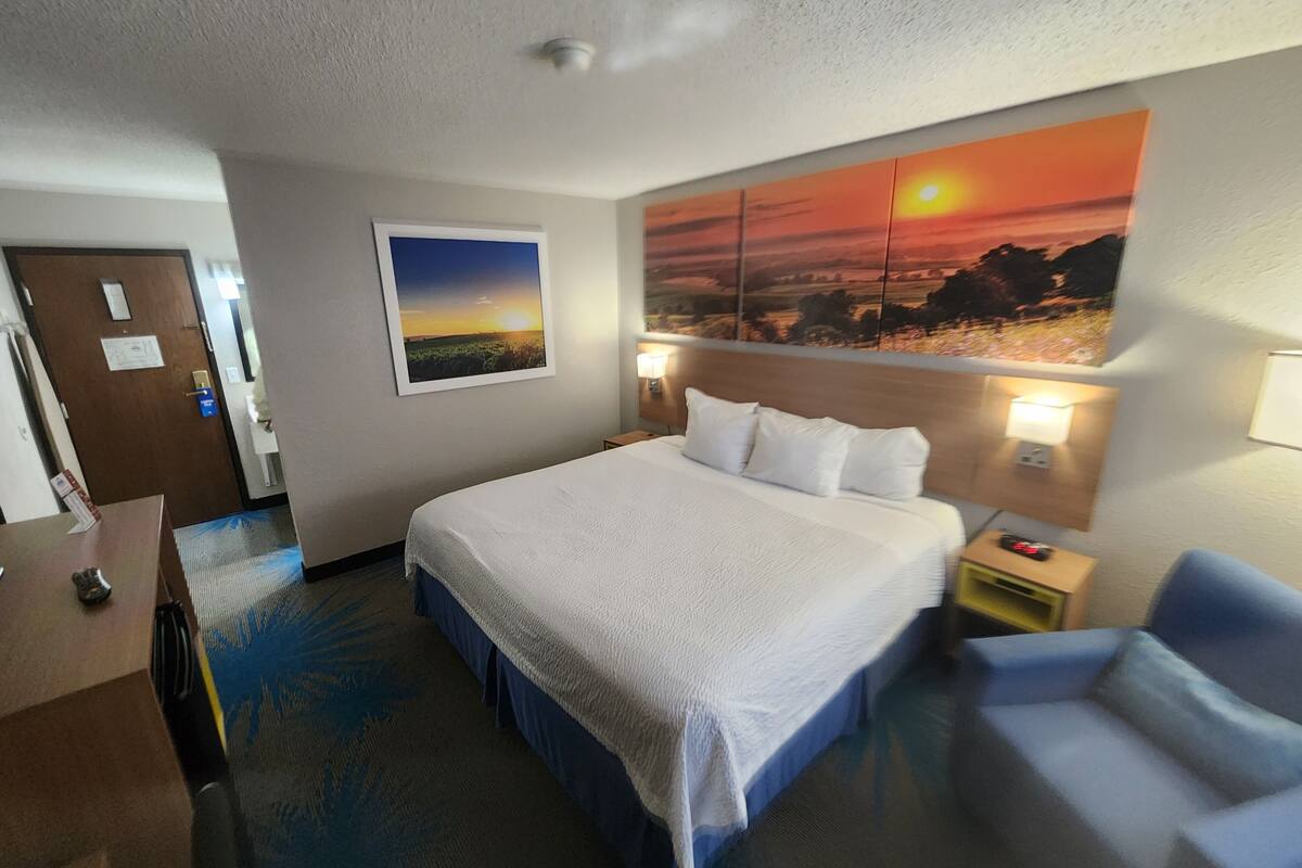 Habitación de hotel con una cama grande, un escritorio, una silla y un cuadro de atardecer en la pared.