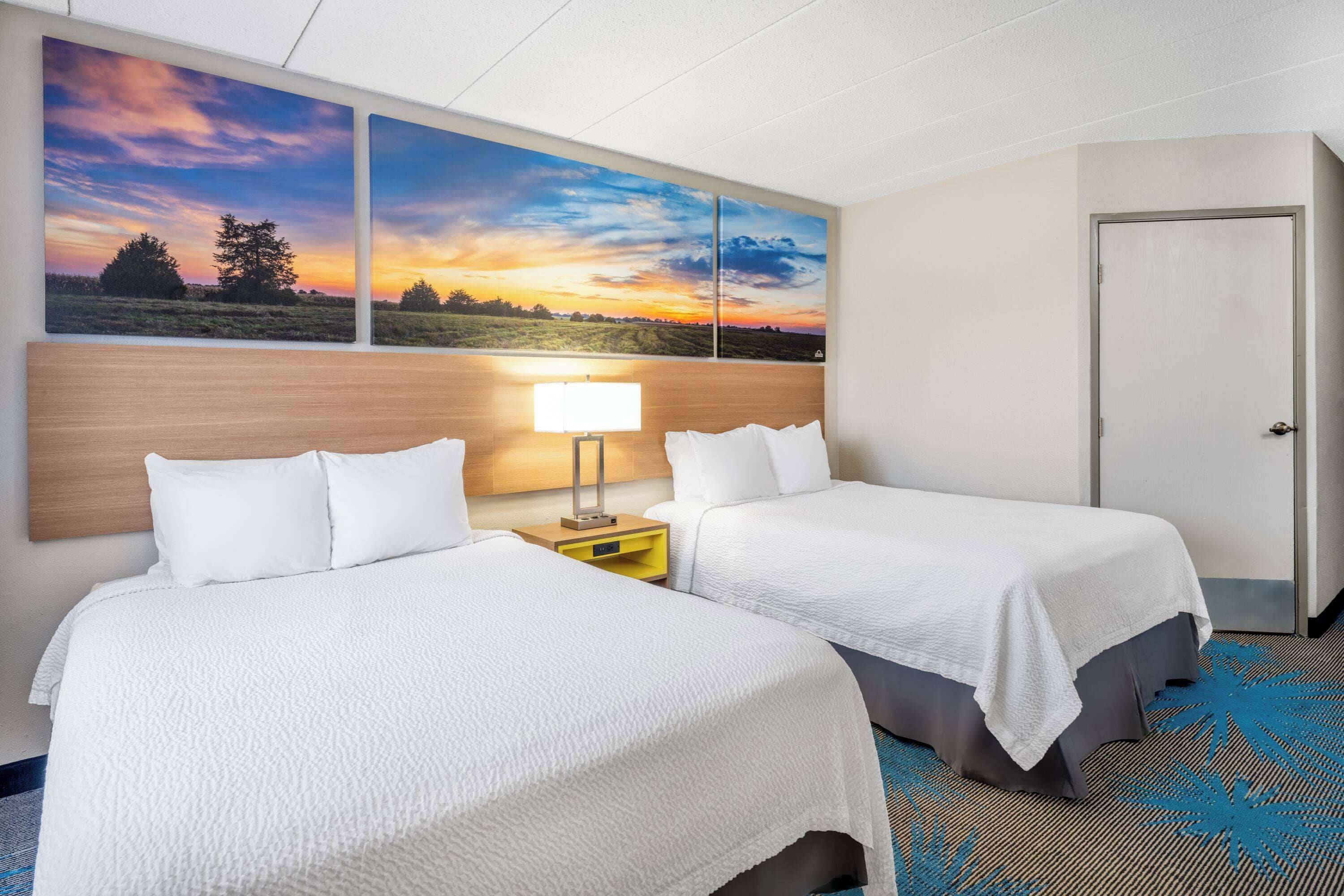 A hotel room with two beds, a nightstand with a lamp, and a large landscape painting on the wall.