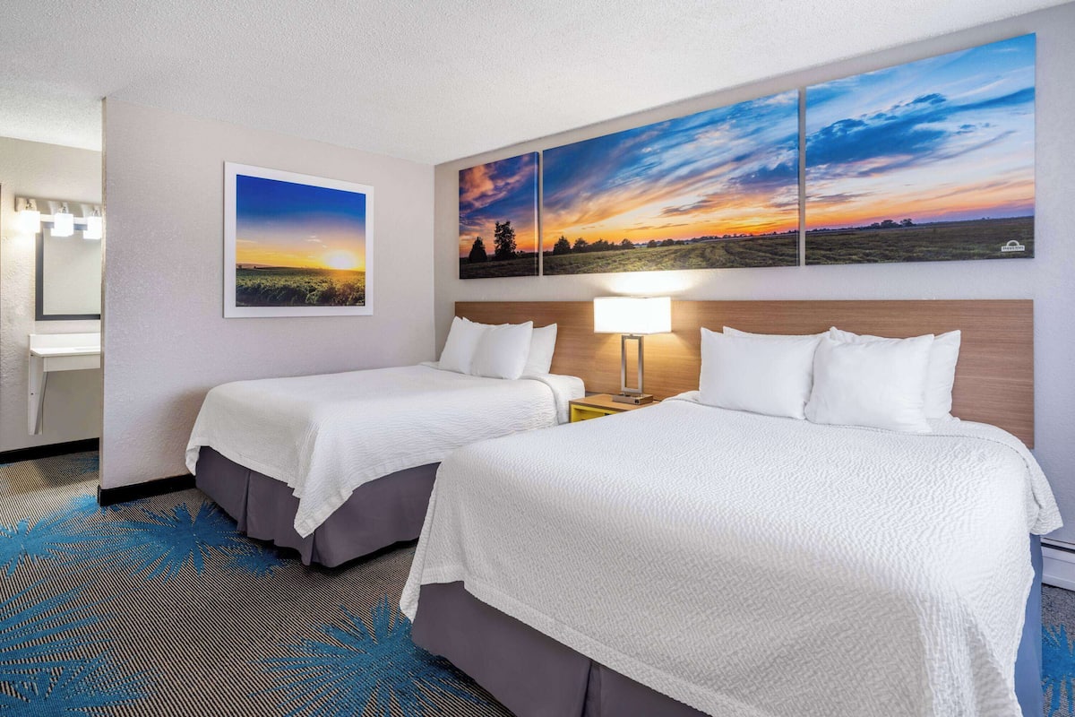 Habitación de hotel con dos camas, una mesita de noche con lámpara, baño con espejo y un cuadro grande con paisaje sobre las camas.