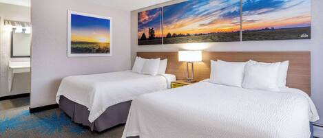 A hotel room with two beds, a nightstand with a lamp, a bathroom with a mirror, and a large landscape painting above the beds.