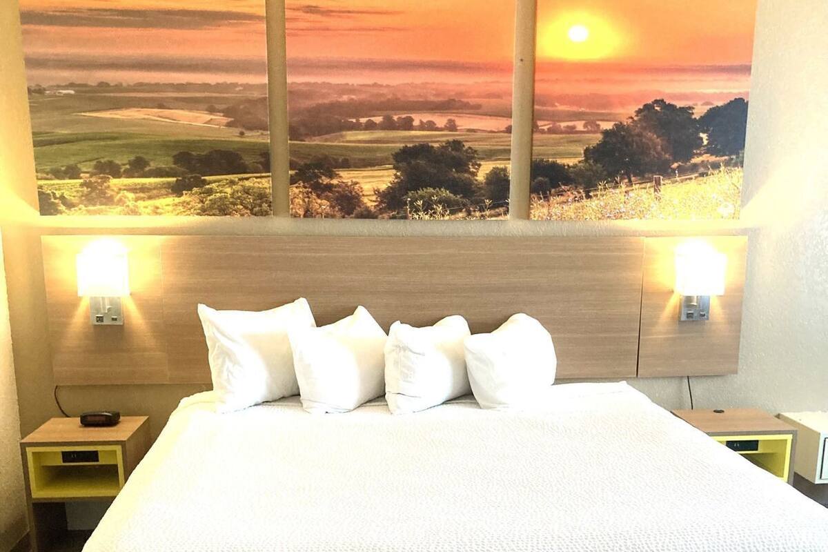 Habitación de hotel con una cama grande, dos lámparas de noche, una luz en la pared y vistas al atardecer.