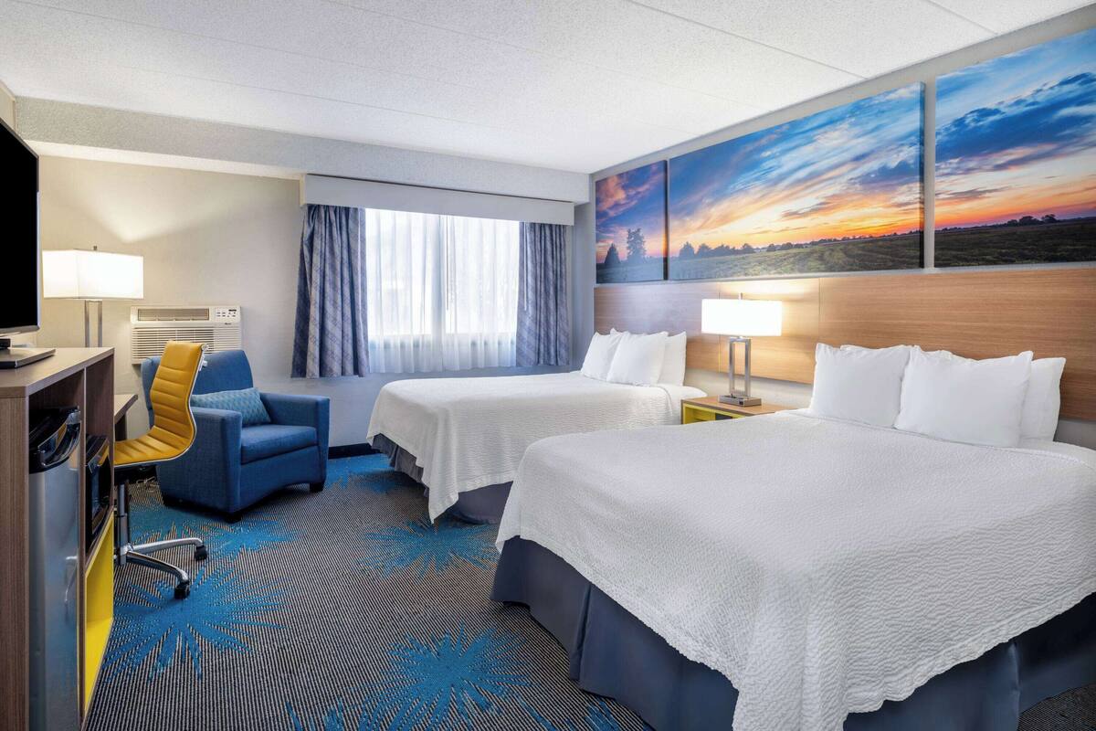 Habitación de hotel con dos camas, un escritorio, una silla y un ventanal grande con cortinas.