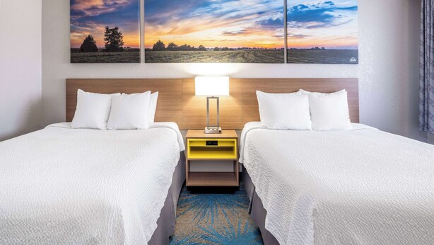 A hotel room with two beds, a nightstand with a lamp, and a large landscape painting above the beds. - Days Inn by Wyndham Sioux Falls Airport (Sioux Falls)