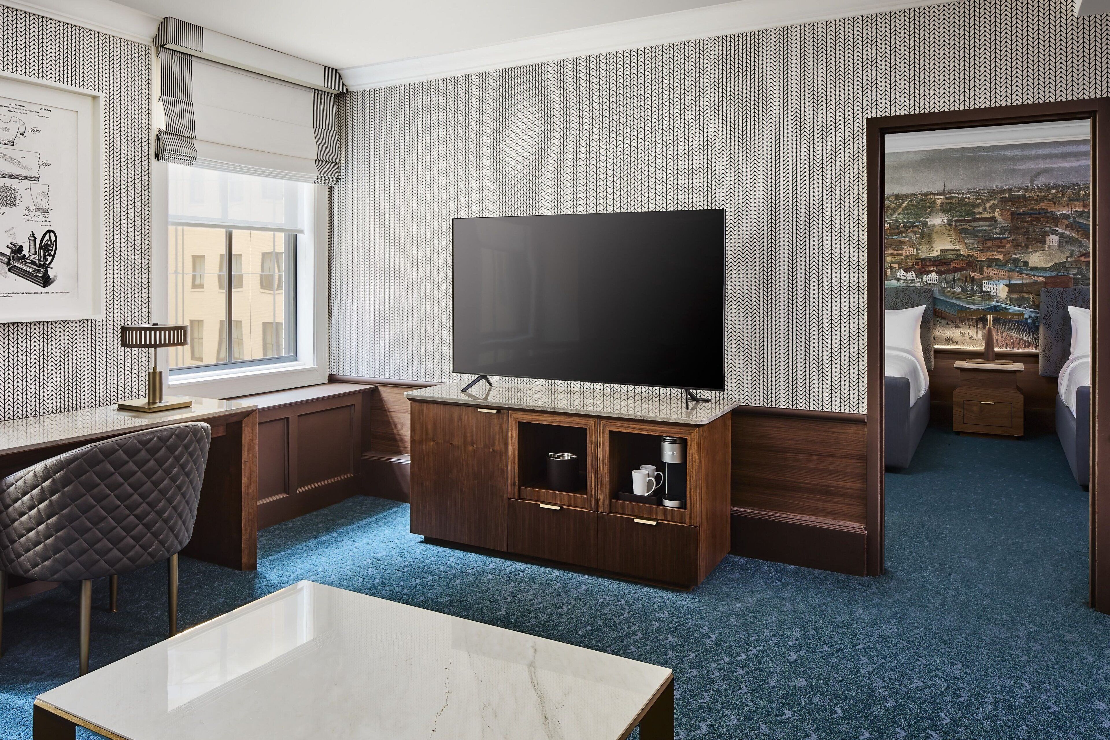 A hotel room with a large flat-screen TV, a wooden cabinet, a desk, a chair, a bed, and a cityscape view through a window.
