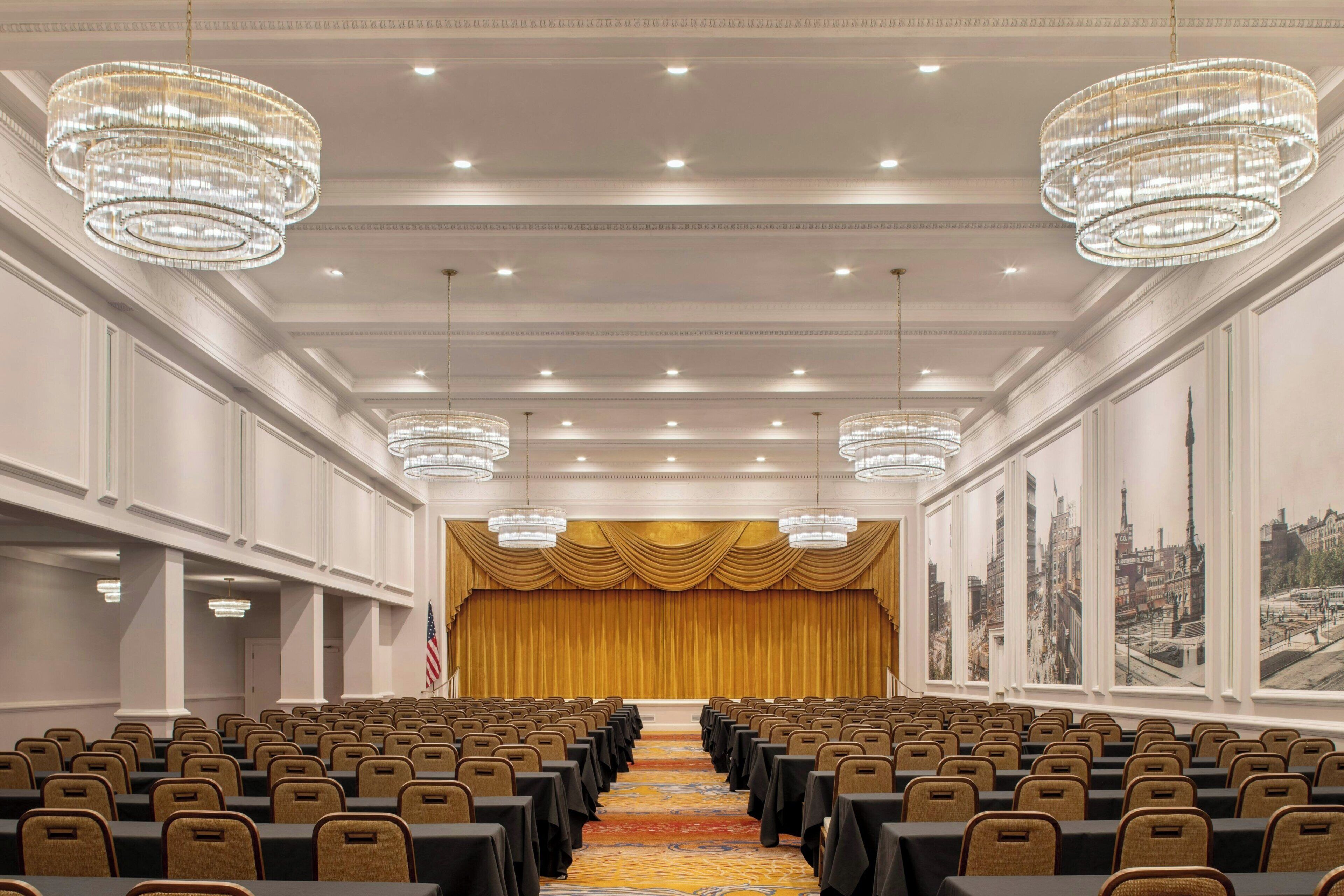 A conference room with rows of chairs, a stage with a golden curtain, and chandeliers.