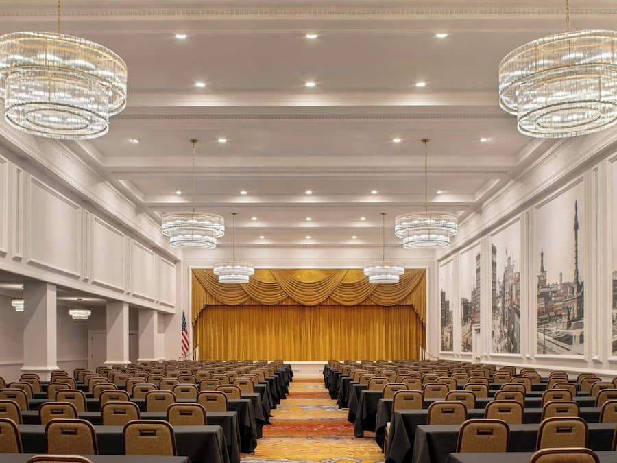 A conference room with rows of chairs, a stage with a golden curtain, and chandeliers.